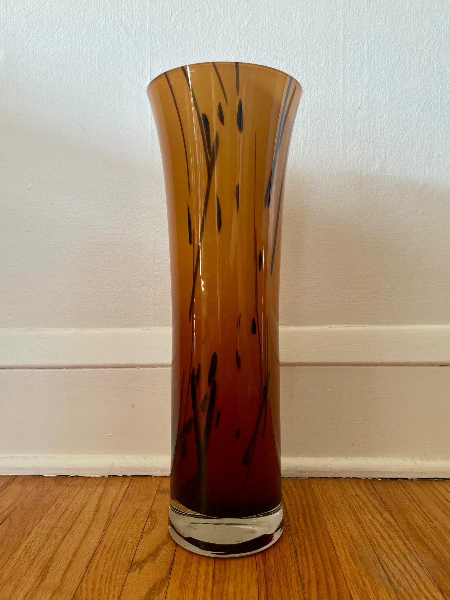 A tall, flared glass vase with a warm amber-to-brown gradient from top to bottom and dark abstract vertical streaks, standing on a wooden floor against a white wall.