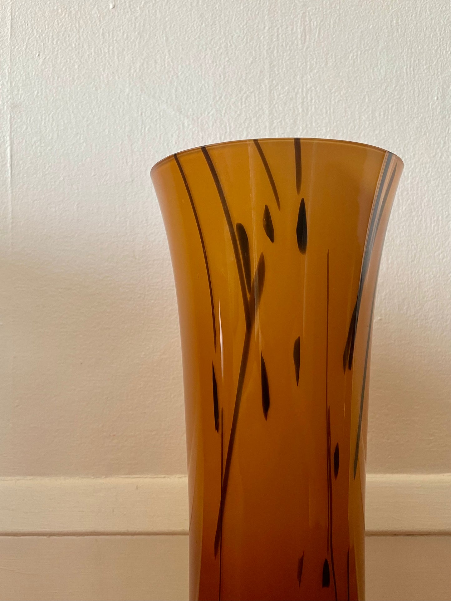 An amber-brown translucent glass vase with a flared rim and black abstract vertical line patterns, shown against a plain off-white wall.