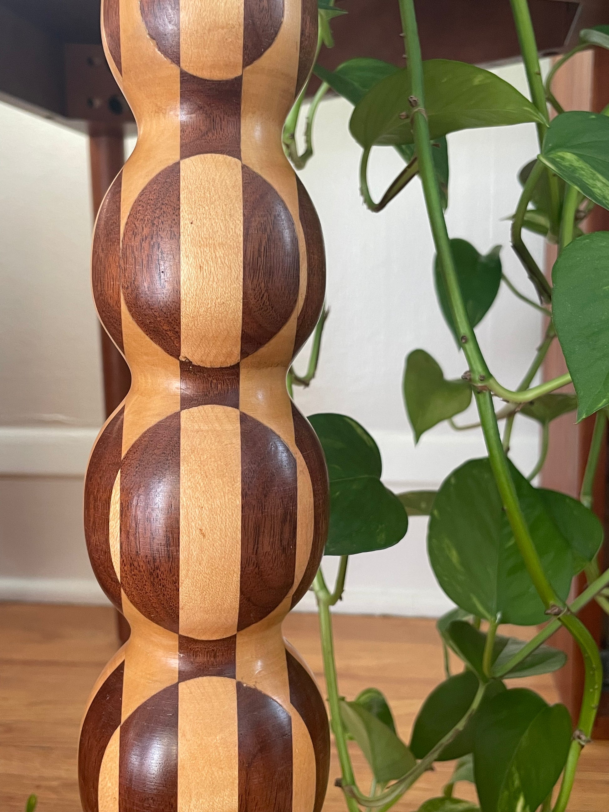 A close-up of a parquetry table lamp featuring a repeating rounded segment pattern in alternating dark and light wood tones, shown indoors next to a green trailing plant.