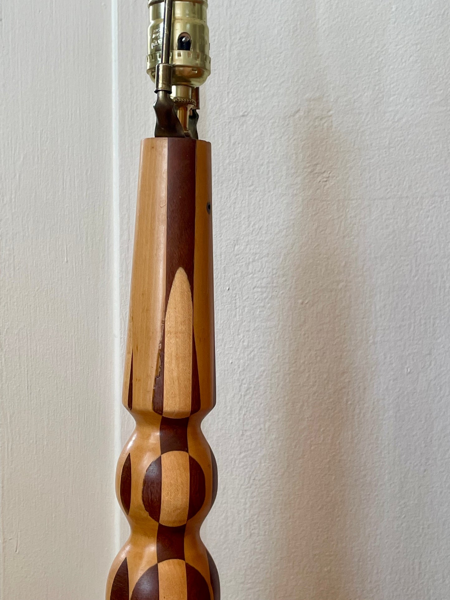 A close-up of a parquetry wooden lamp base featuring a pattern with alternating light and dark wood, with a brass-colored lamp socket at the top, set against a plain off-white wall.