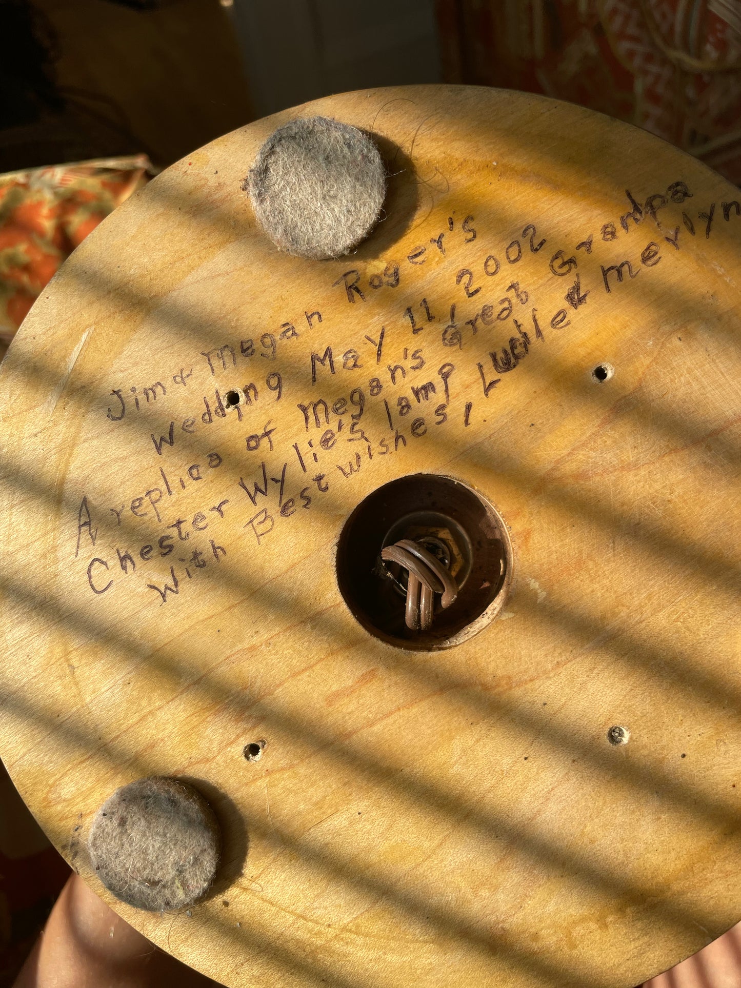A close-up of the underside of a wood lamp with handwritten text  and two round gray felt pads. Strong diagonal sunlight casts shadows across the wood, and a hand is partially visible holding the object.