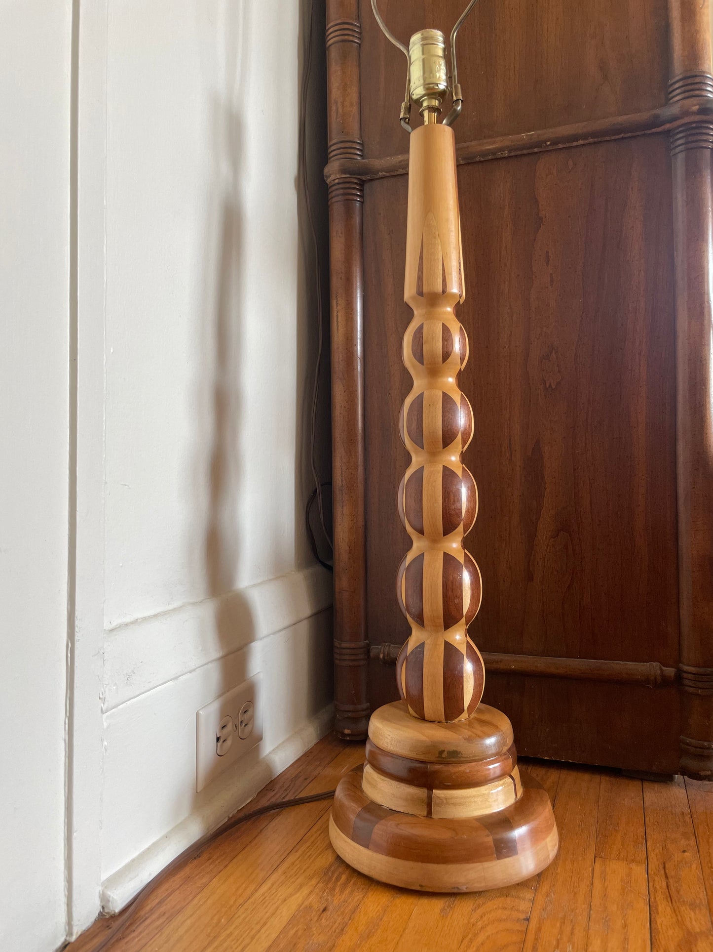 A handmade wooden table lamp with a decorative turned spindle column and tiered base, shown on a wooden floor next to a white wall and dark wood cabinet.