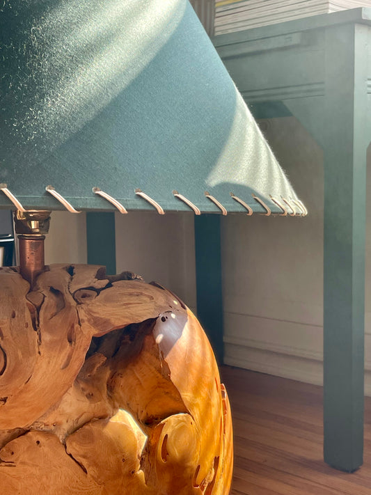 A close-up of a round polished wood lamp showcasing the natural variation and organic shape of a root ball, with a copper neck and a large dark green fabric shade with whipstitching around the edge and curved rays of light falling across the piece, set in an indoor room with a green painted table in the background.