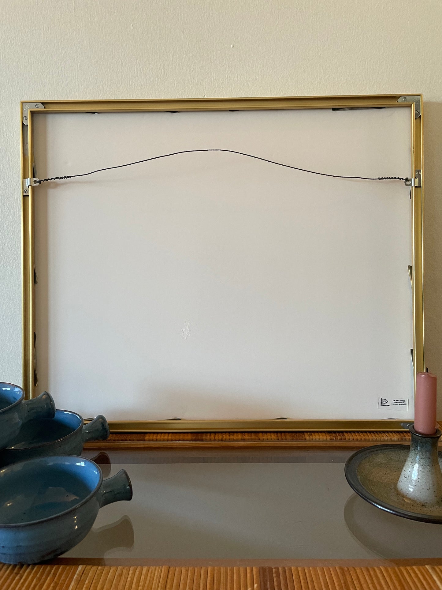 Backside of a picture frame with a gold-toned metal border and a hanging wire at the top, displayed against a beige wall with decorative bowls and a candle on a table below.