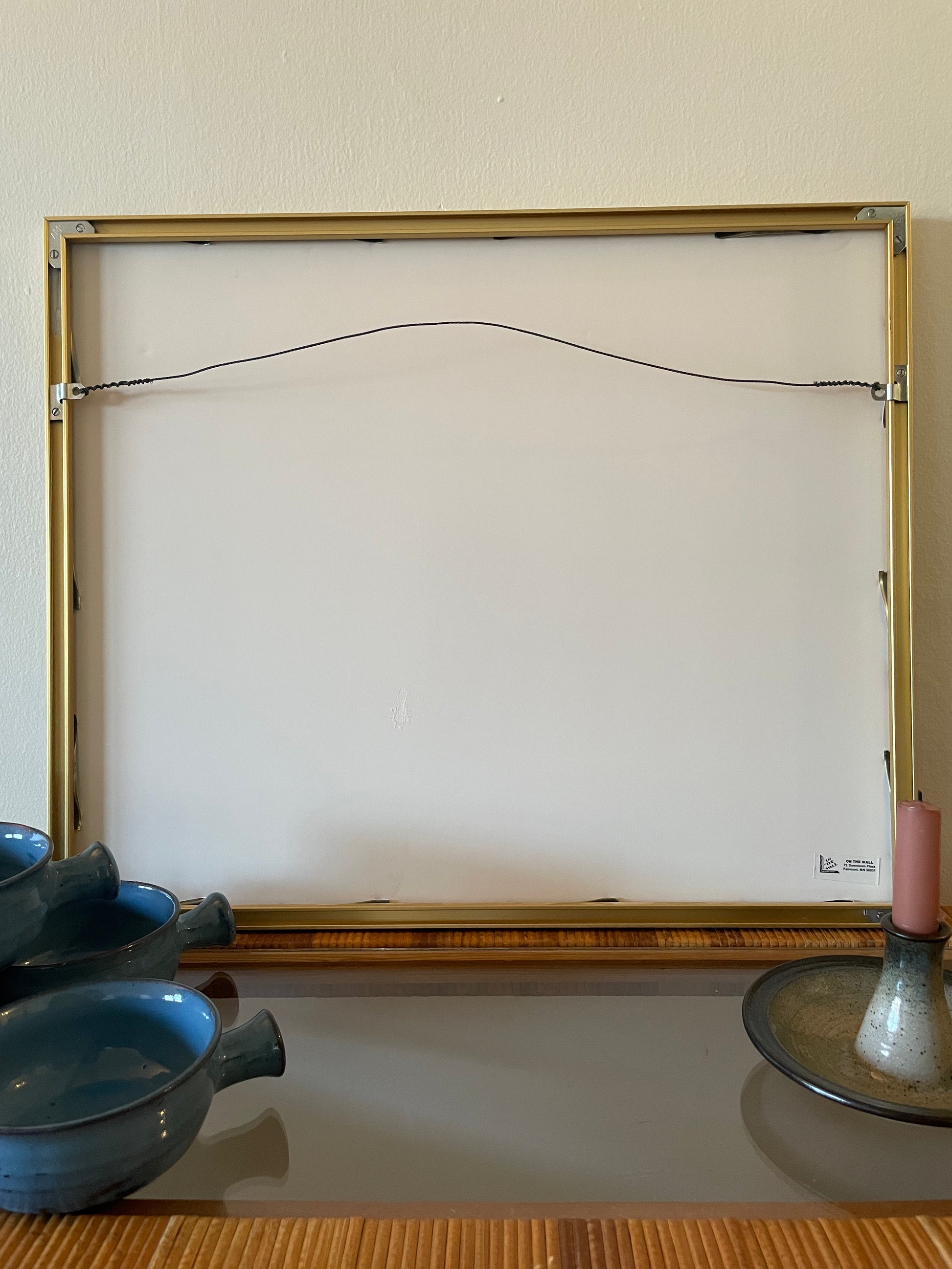 Backside of a picture frame with a gold-toned metal border and a hanging wire at the top, displayed against a beige wall with decorative bowls and a candle on a table below.