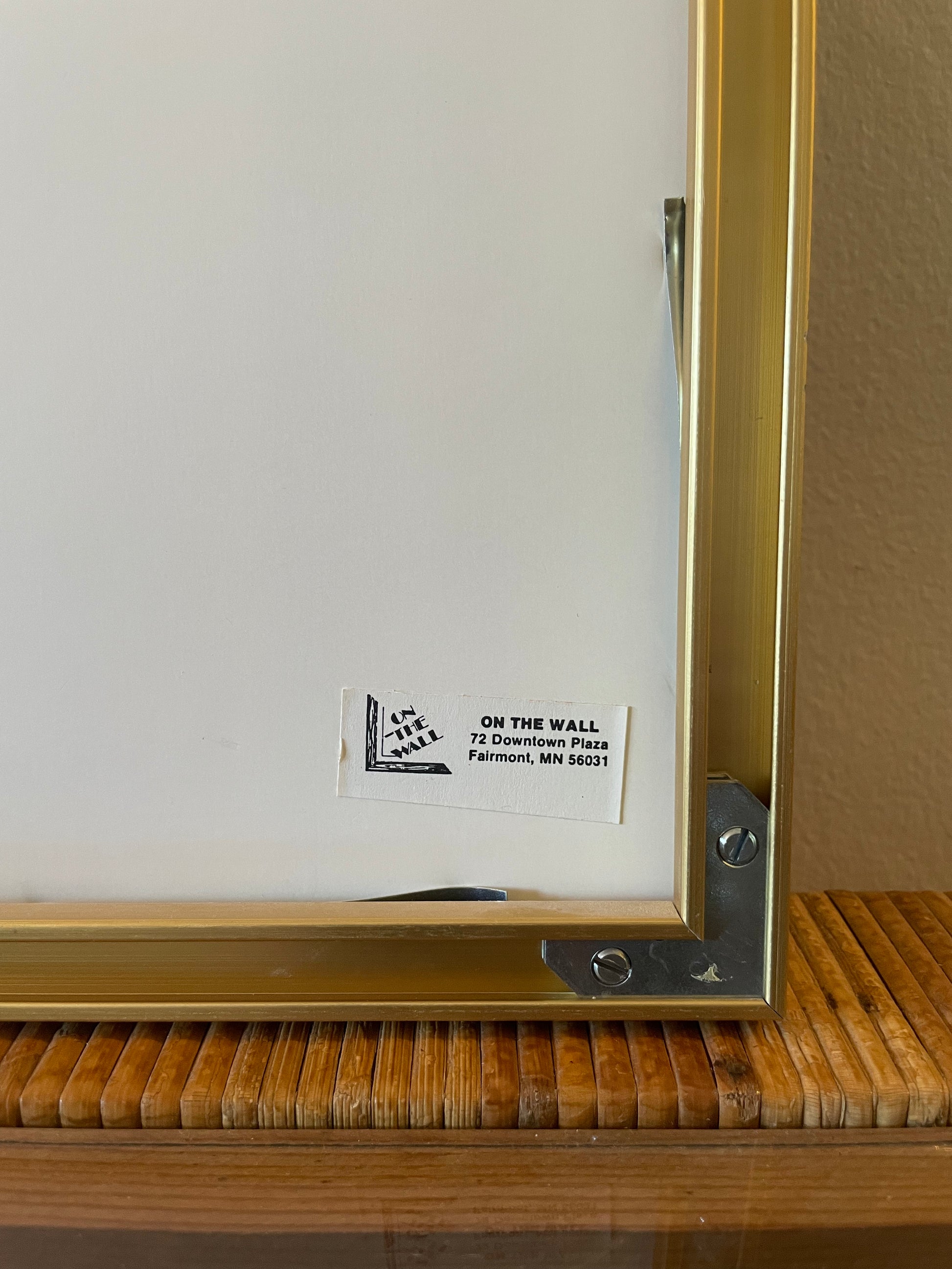Close-up of the back corner of framed wall art with a gold frame, metal mounting bracket, and a small seller label reading 'ON THE WALL'.