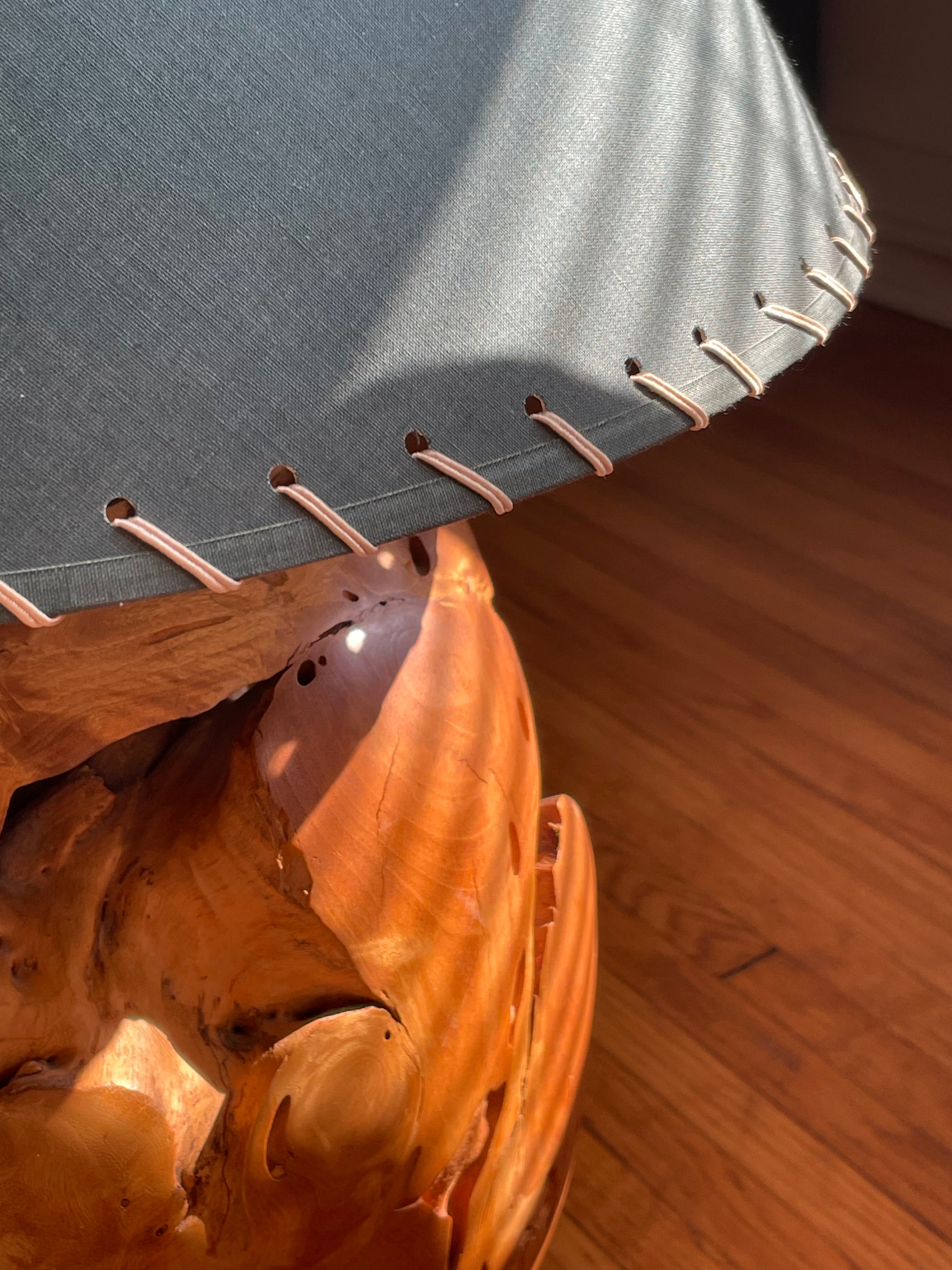 A close-up of an organically-shaped round wooden lamp base with a dark green fabric lampshade featuring whipstitched edges, set on a polished hardwood floor.