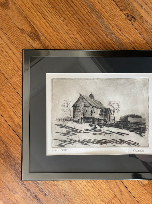 A framed black and white artist's state proof of a rustic barn and silo landscape signed ‘Rozanski', displayed on a warm brown wooden floor. The artwork is enclosed in a glossy black frame with a black mat border.