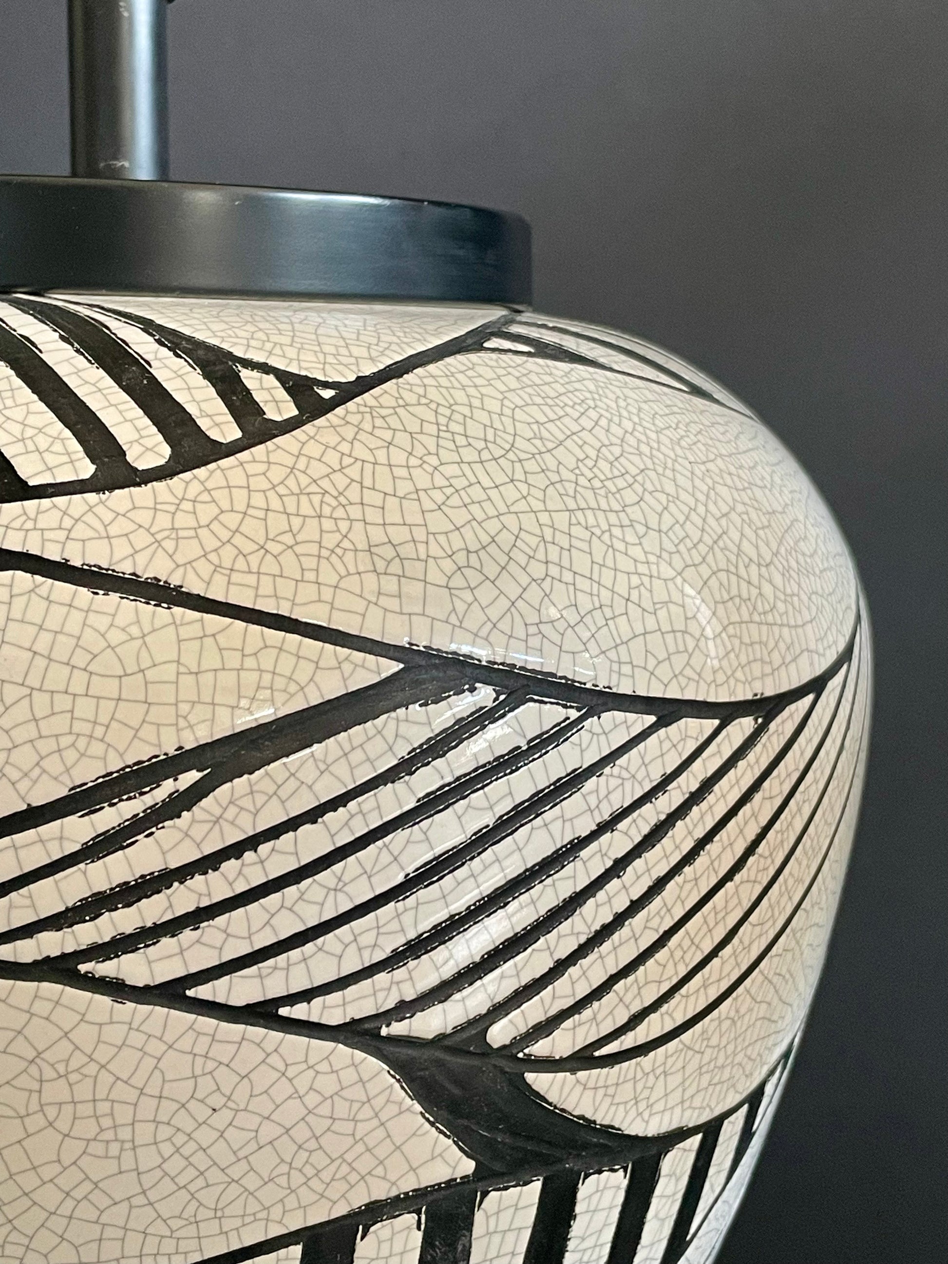 Close-up of a ceramic table lamp with cream crackle glaze and black painted leaf-like geometric pattern in relief, and a dark metal top against a dark gray background.