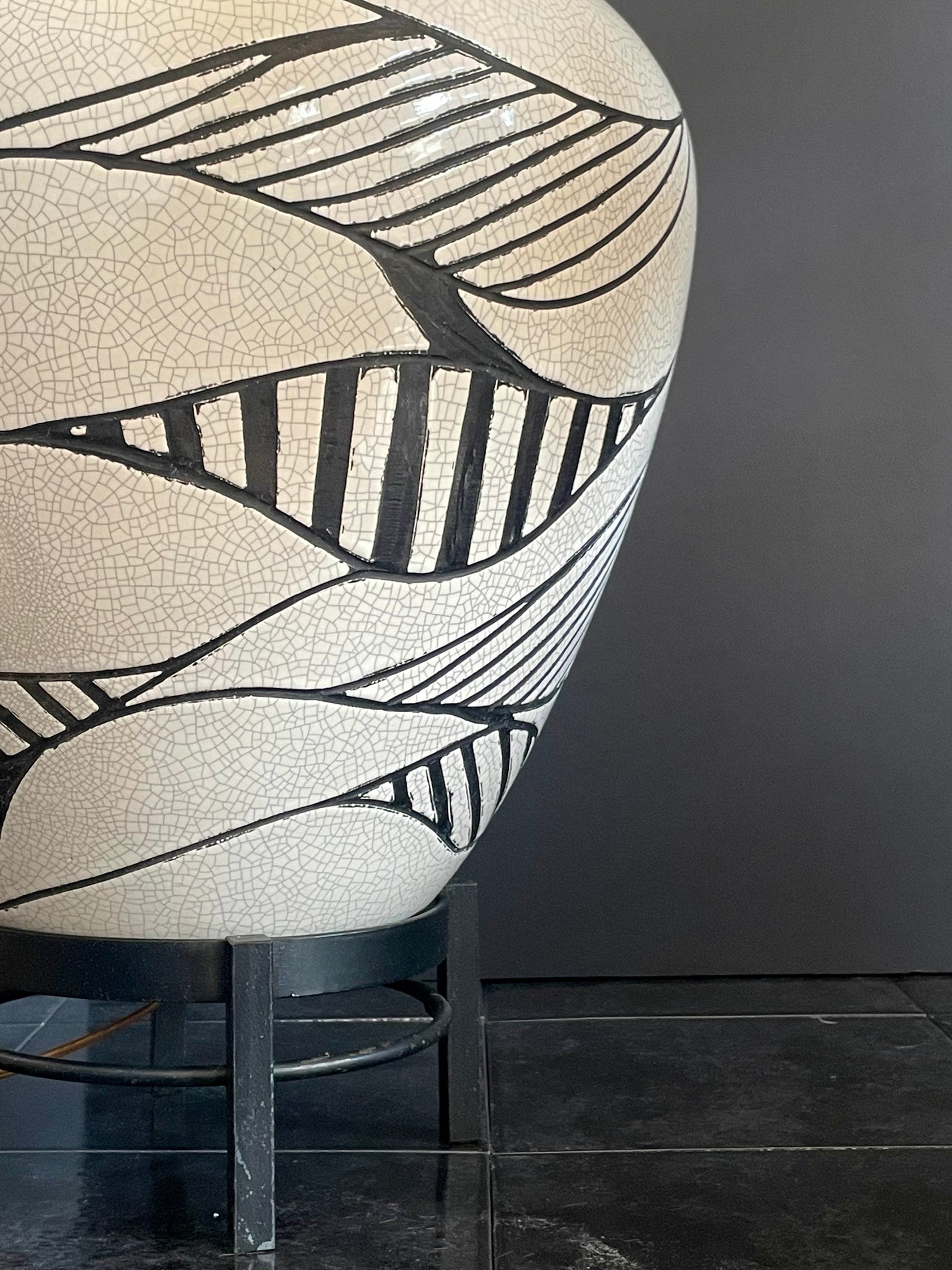 A close-up of a large ceramic lamp base with a cream-colored crackle glaze and bold black leaf-like line patterns in relief, displayed on a black metal stand against a dark gray wall and black tiled floor.