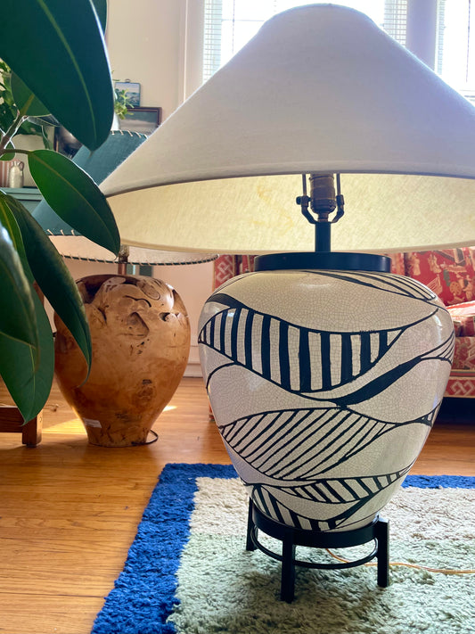 A ceramic table lamp with a black and white geometric patterned base and an off-white linen shade, set on a black metal stand on a blue and white shag rug in a living room.