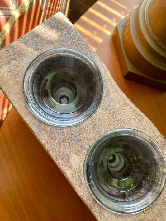 A partial view of a rustic rectangular wooden block showing two of three inset circular glass votives, displayed on a wooden table in warm natural light.