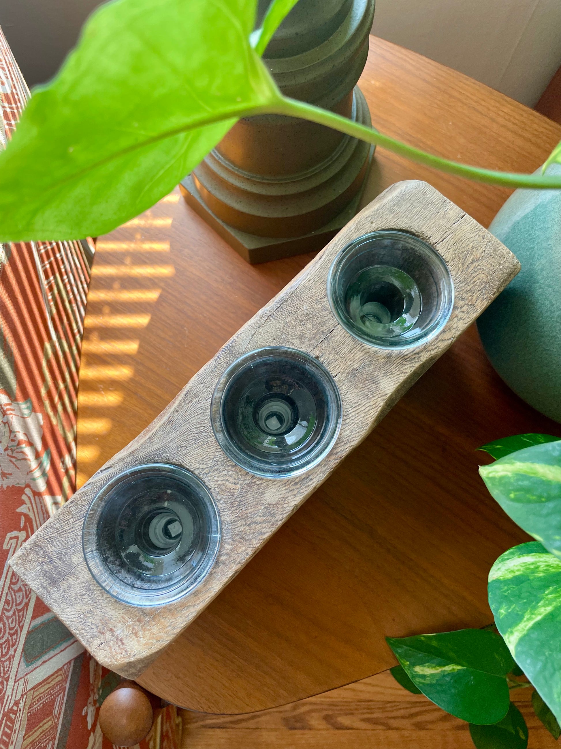 A rustic rectangular wooden block with three inset circular glass votives, displayed on a wooden table with green plants in a home setting.