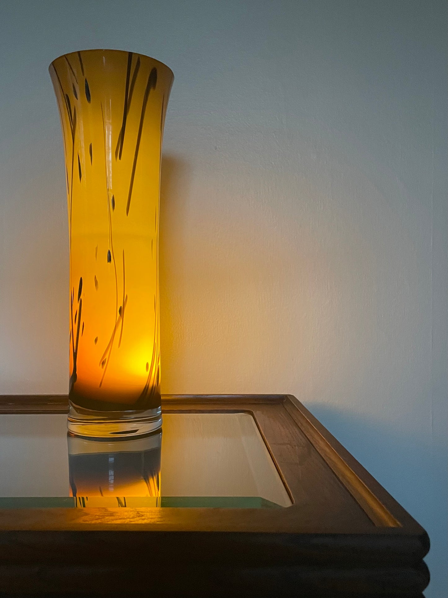 A tall, flared amber glass vase with black abstract streaks, illuminated from within to glow warm yellow-orange, set on a glass-topped wooden table against a plain wall.