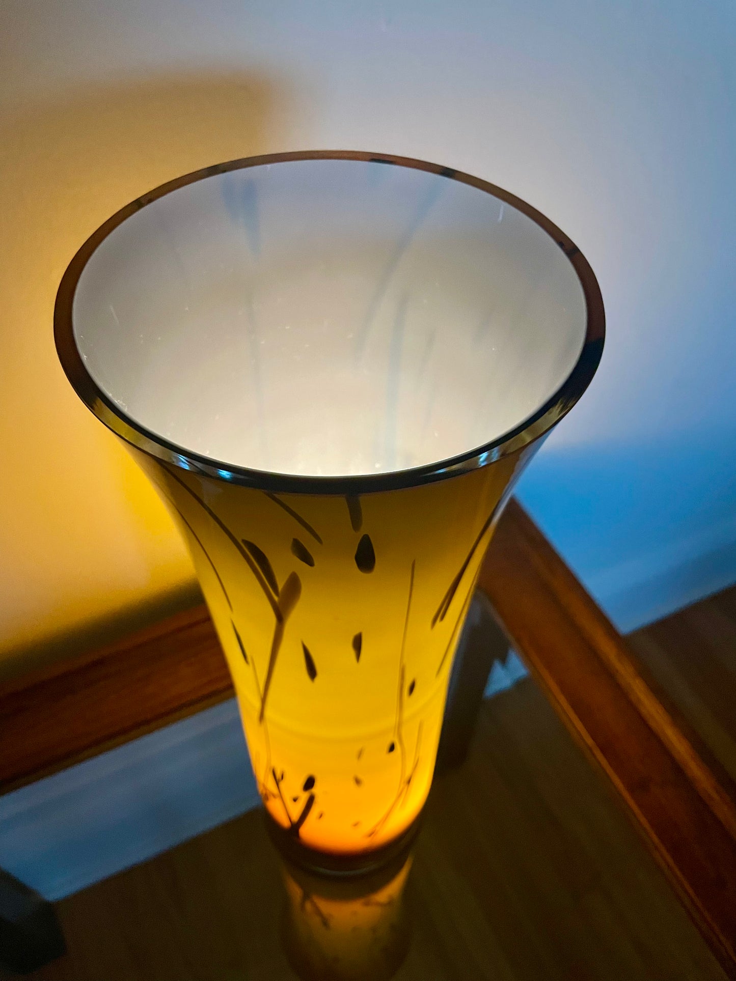 A tall, flared glass vase with a warm yellow-orange glow from within, featuring black leaf-like etched patterns on its exterior. The vase is photographed from above on a glass-top wood table against a white wall with a blue light reflection.