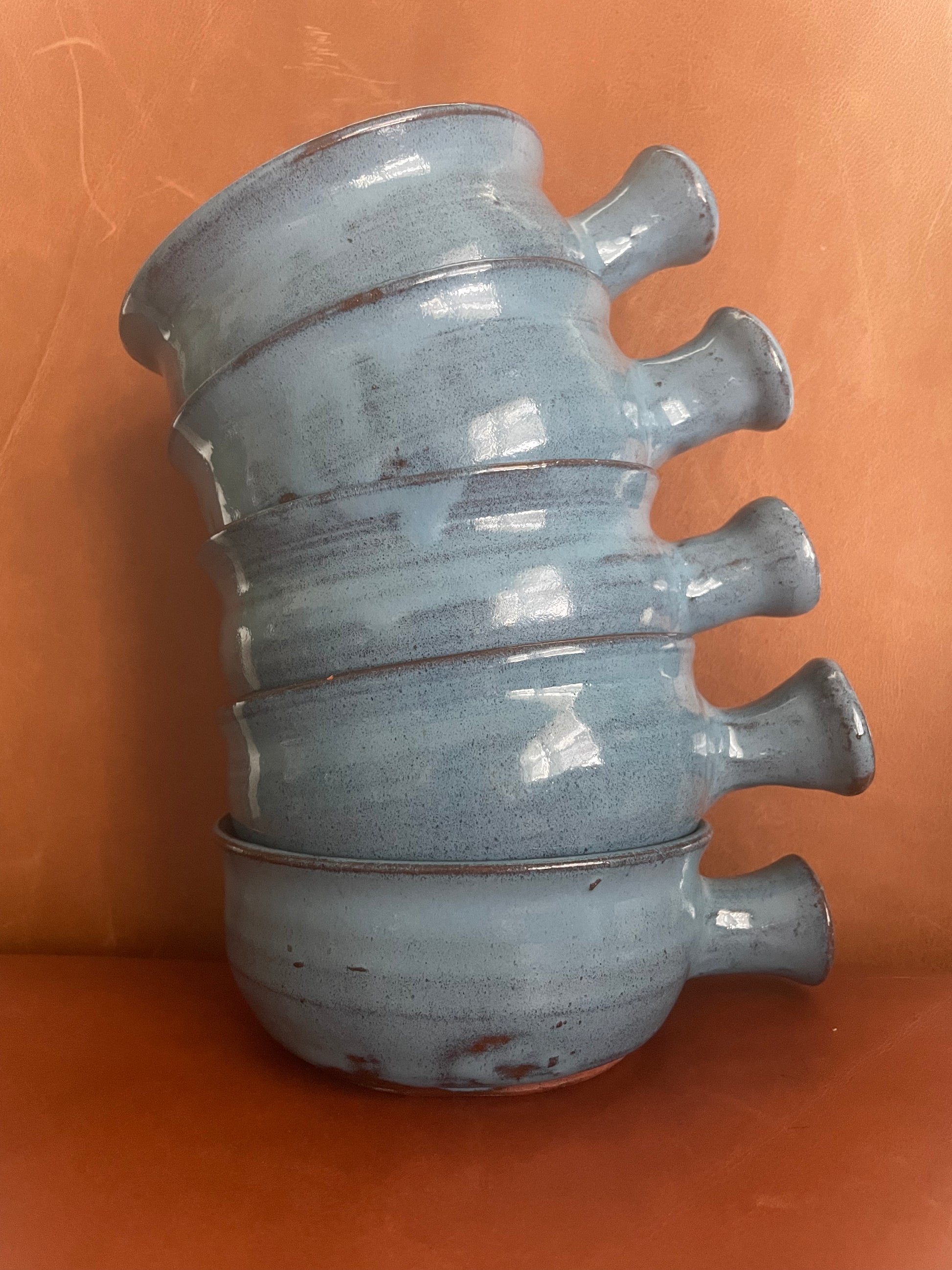 A stack of five handmade ceramic stoneware bowls with side handles, glazed in an irregular light blue-gray color over terracotta clay, set against a warm terracotta background.