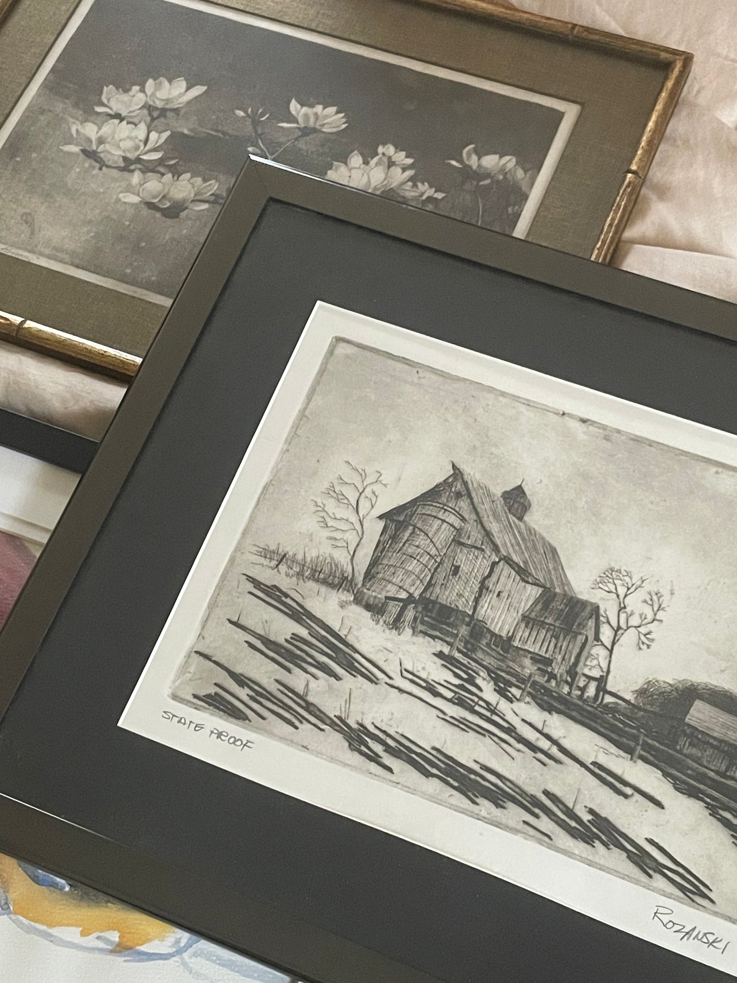 A framed monochrome art print showing a rustic farm landscape, photographed alongside other framed artworks on a bed.