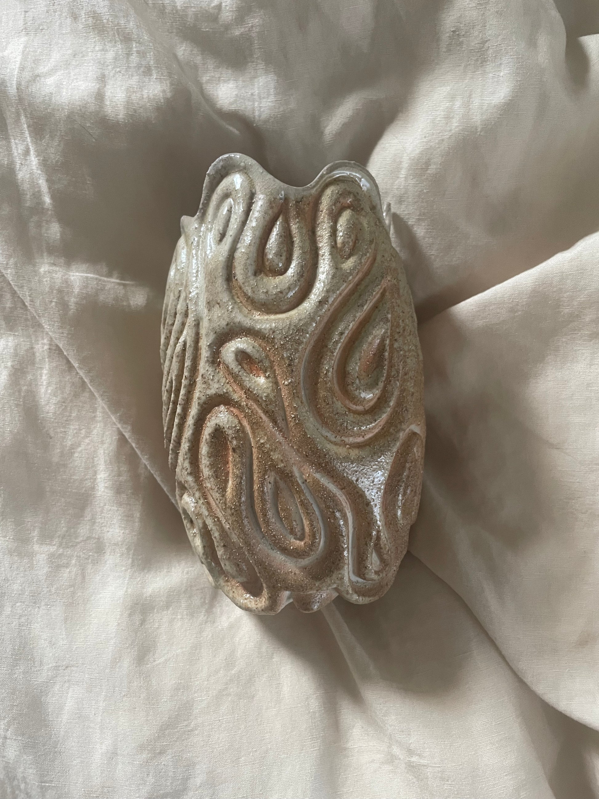 An organic-shaped ceramic vase with a textured, swirling pattern, resting on draped off-white linen.
