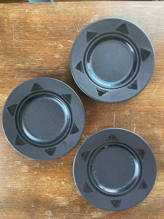 Three round black ceramic plates with a raised rim and small triangular pattern around the rim, arranged on a wooden tabletop.