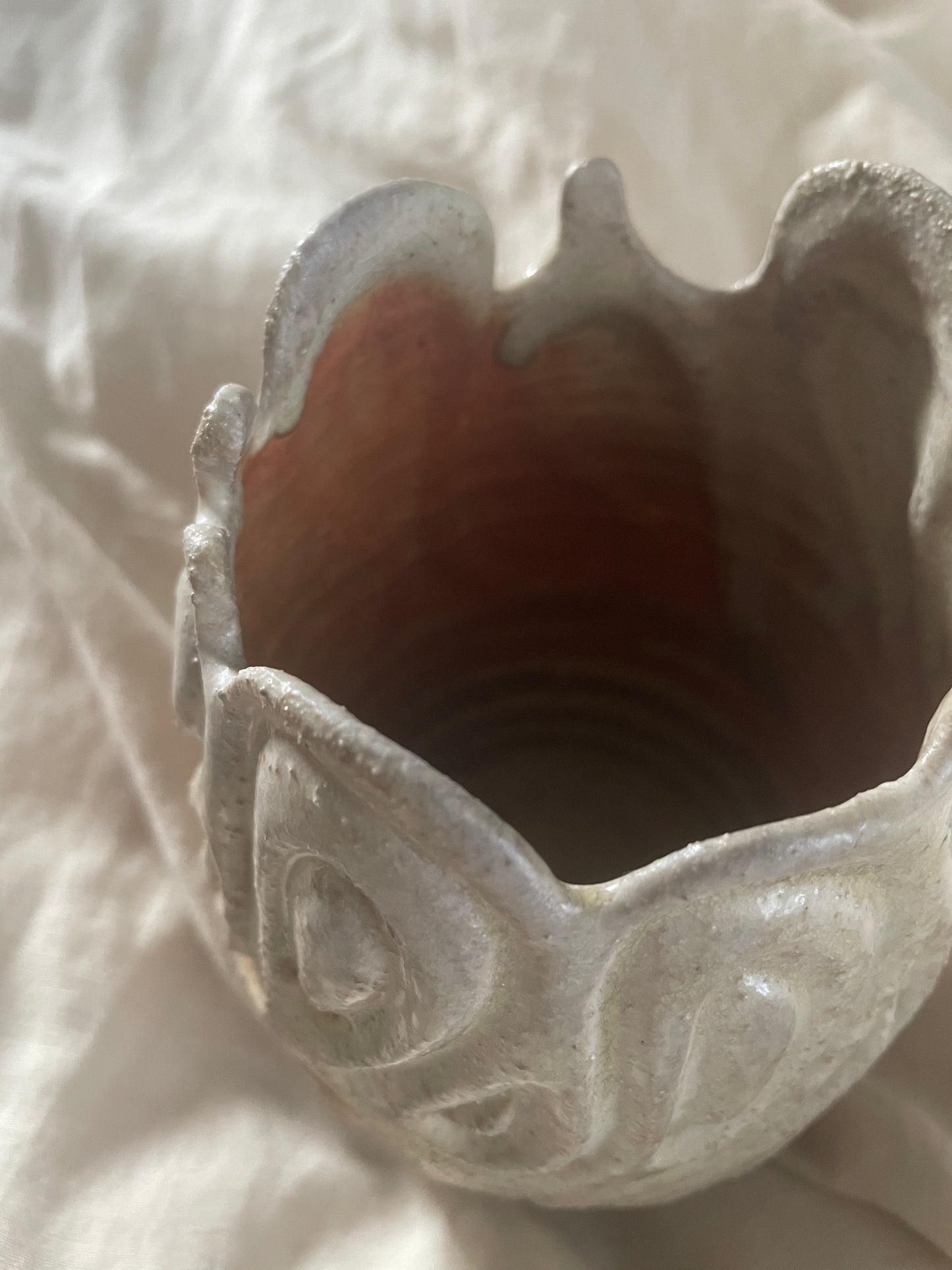 A handmade ceramic vase with a textured, organic scalloped rim and carved abstract swirl patterns, shown from above on a draped off-white linen background.