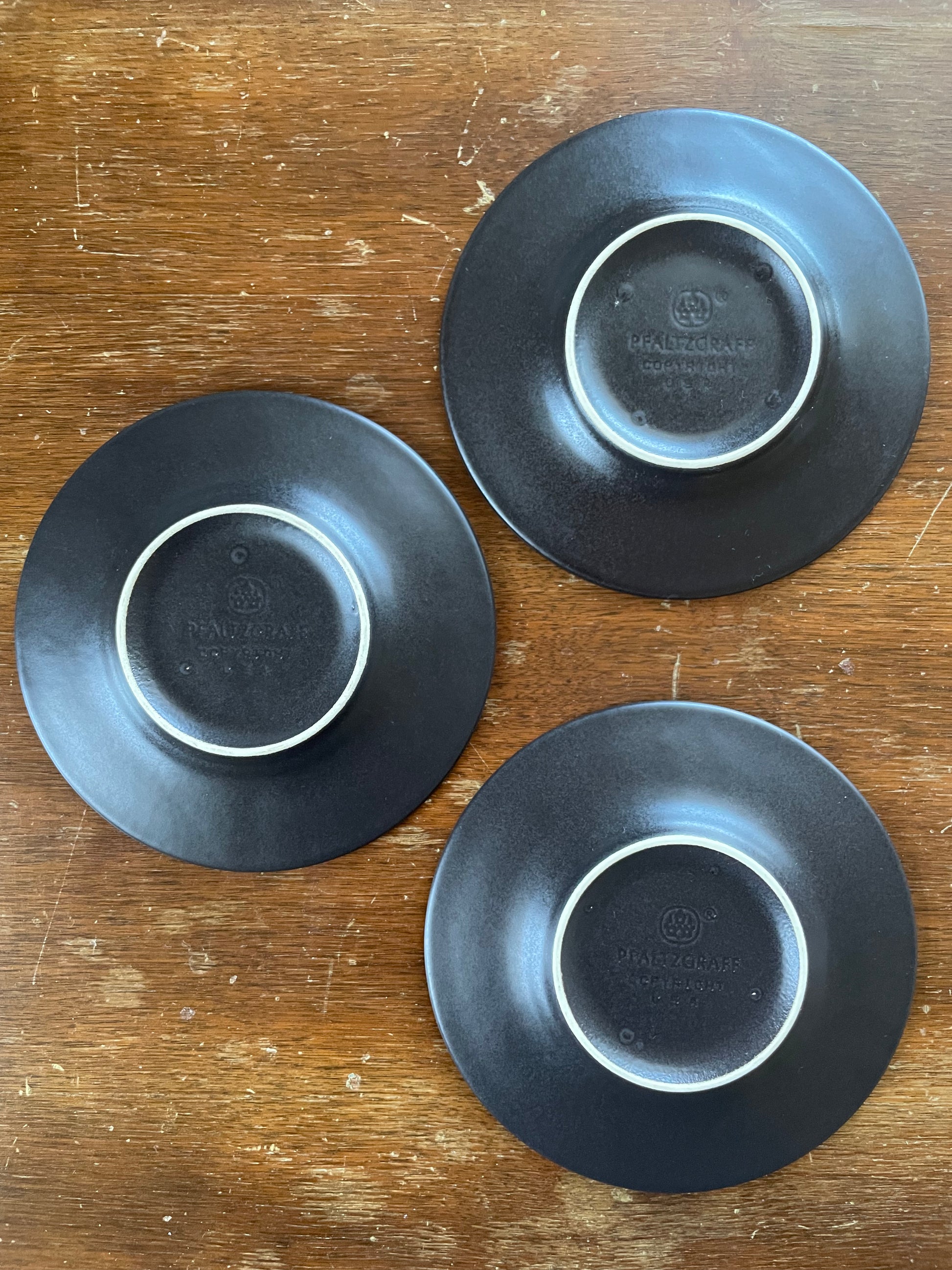 Three black ceramic plates shown from the bottom side, revealing a white rim and a circular embossed logo that reads ‘Pfaltzgraff', placed on a wooden surface.