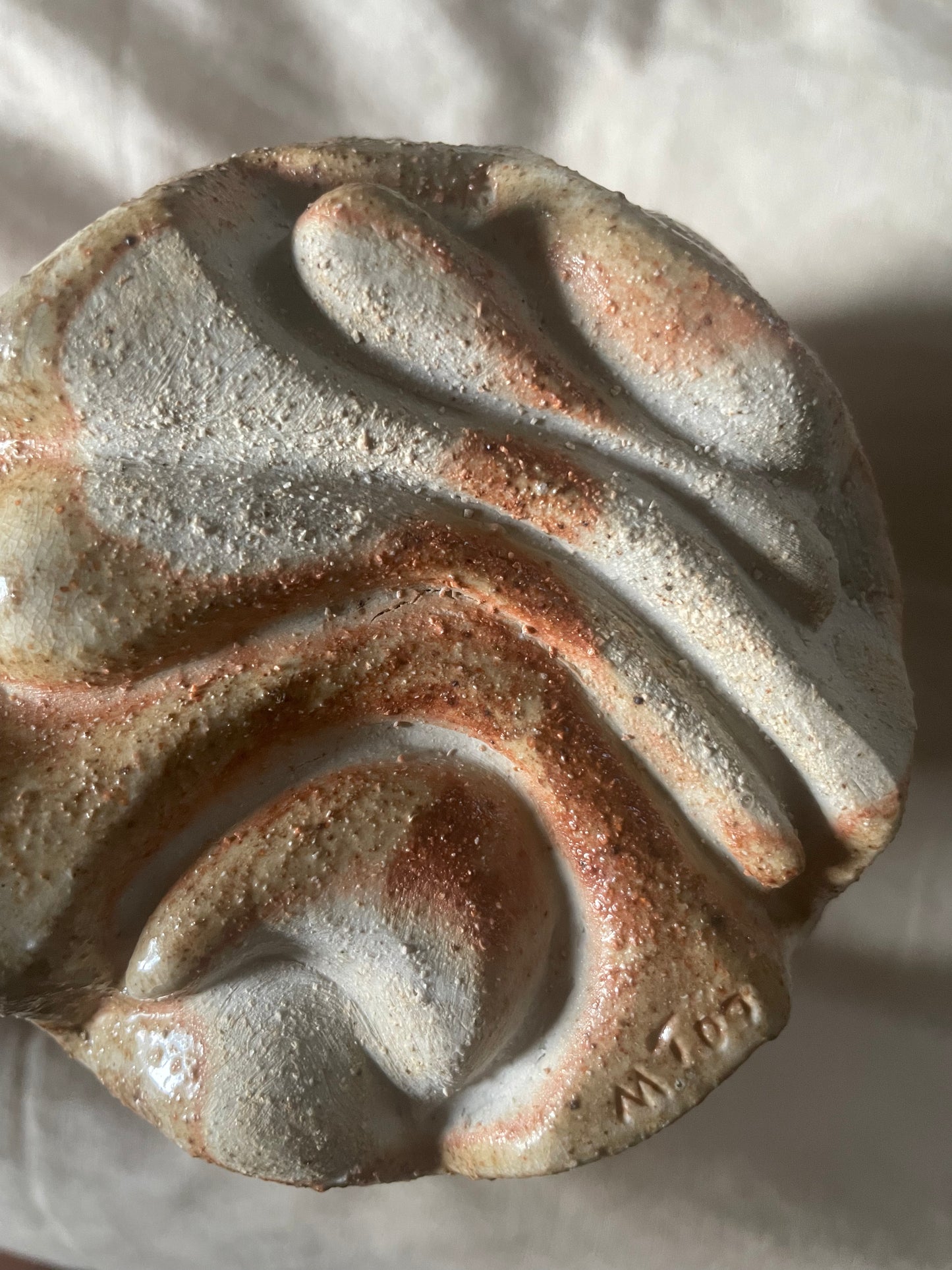 Close-up of the round underside of a ceramic vase with a carved organic swirly pattern in toasty beige and terracotta glazes, resting on an off-white linen background.