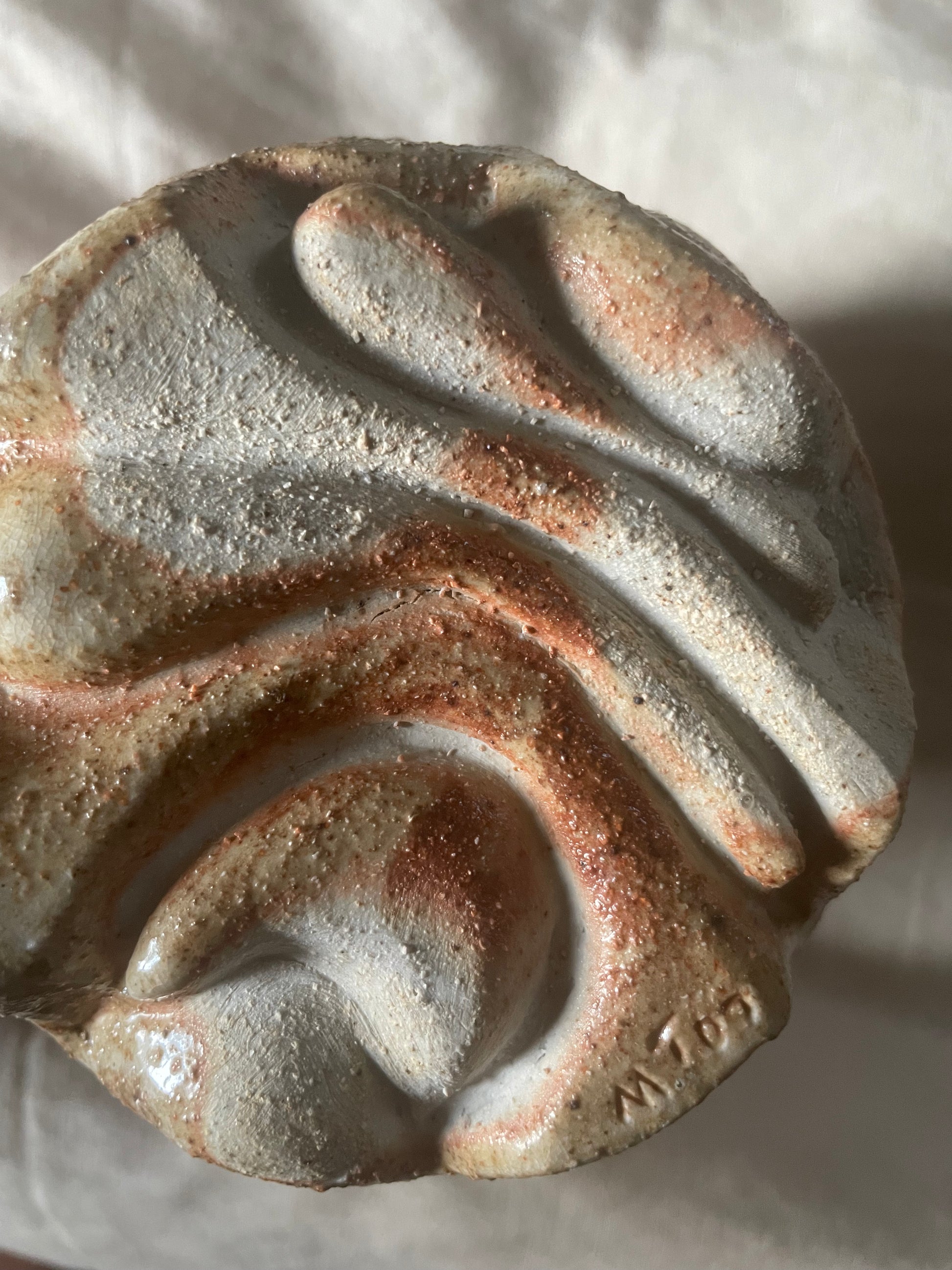 Close-up of the round underside of a ceramic vase with a carved organic swirly pattern in toasty beige and terracotta glazes, resting on an off-white linen background.