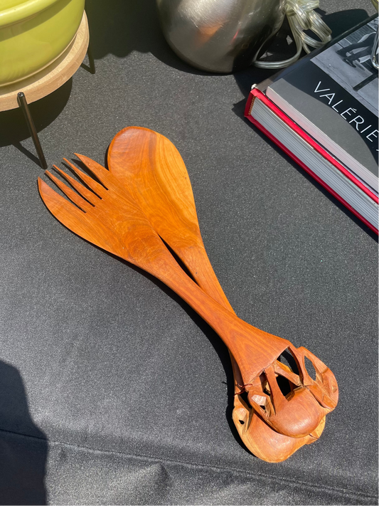 A spoon and forked salad server with elephants carved at the top rests on a black surface with books and other objects shown at the edges of the image.