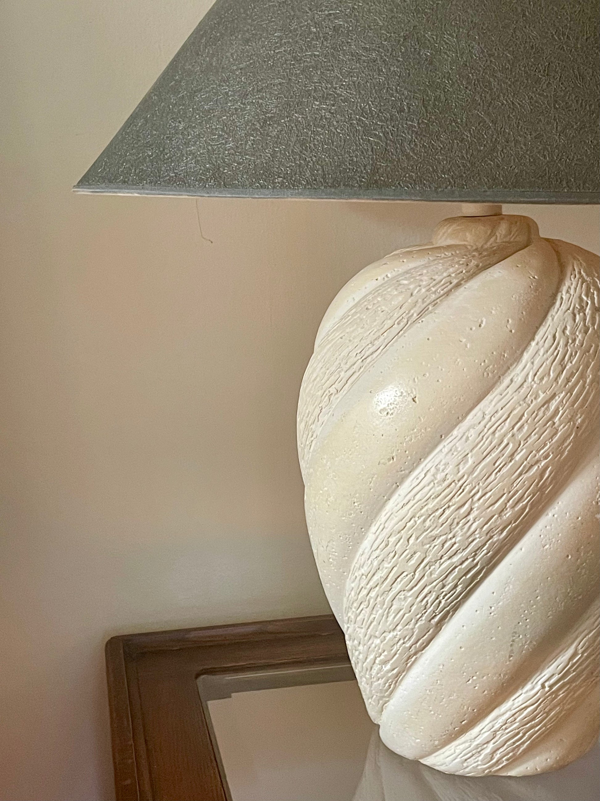 A close-up of a cream-colored plaster table lamp base with a textured spiral design shown with a graphite tapered lampshade, set against a warm beige wall.