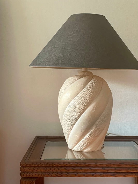 A cream-colored plaster table lamp base with a textured spiral design shown with a graphite tapered lampshade, sitting on a glass-top wood side table in front of a plain wall.