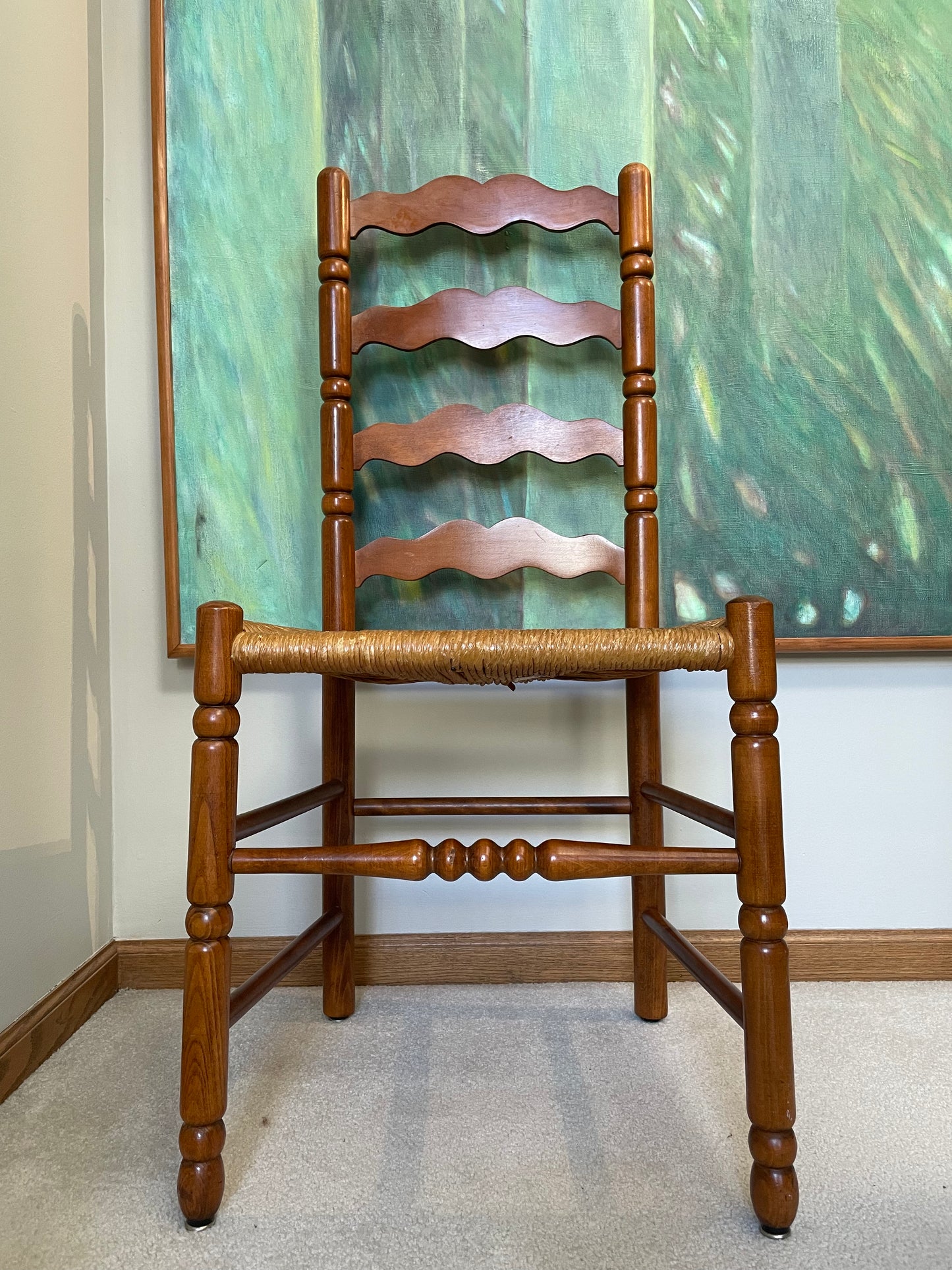 A wooden ladder-back dining chair with a woven rush seat, positioned in front of a green abstract painting on a wall.