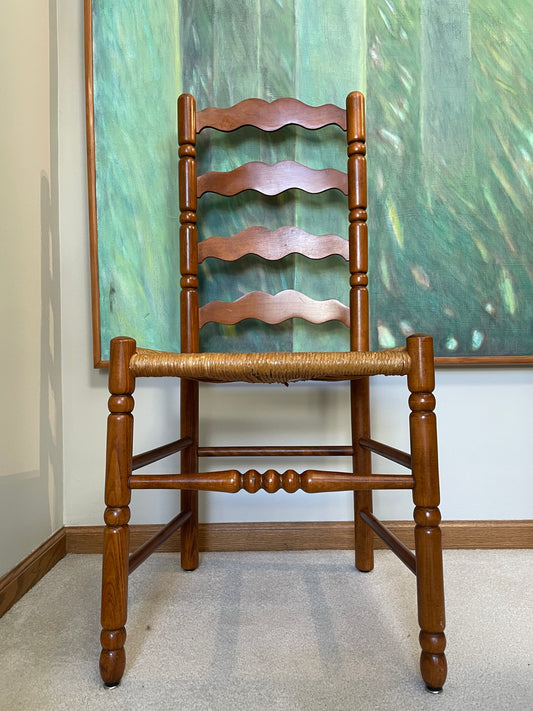 A wooden ladder-back dining chair with a woven rush seat, positioned in front of a green abstract painting on a wall.