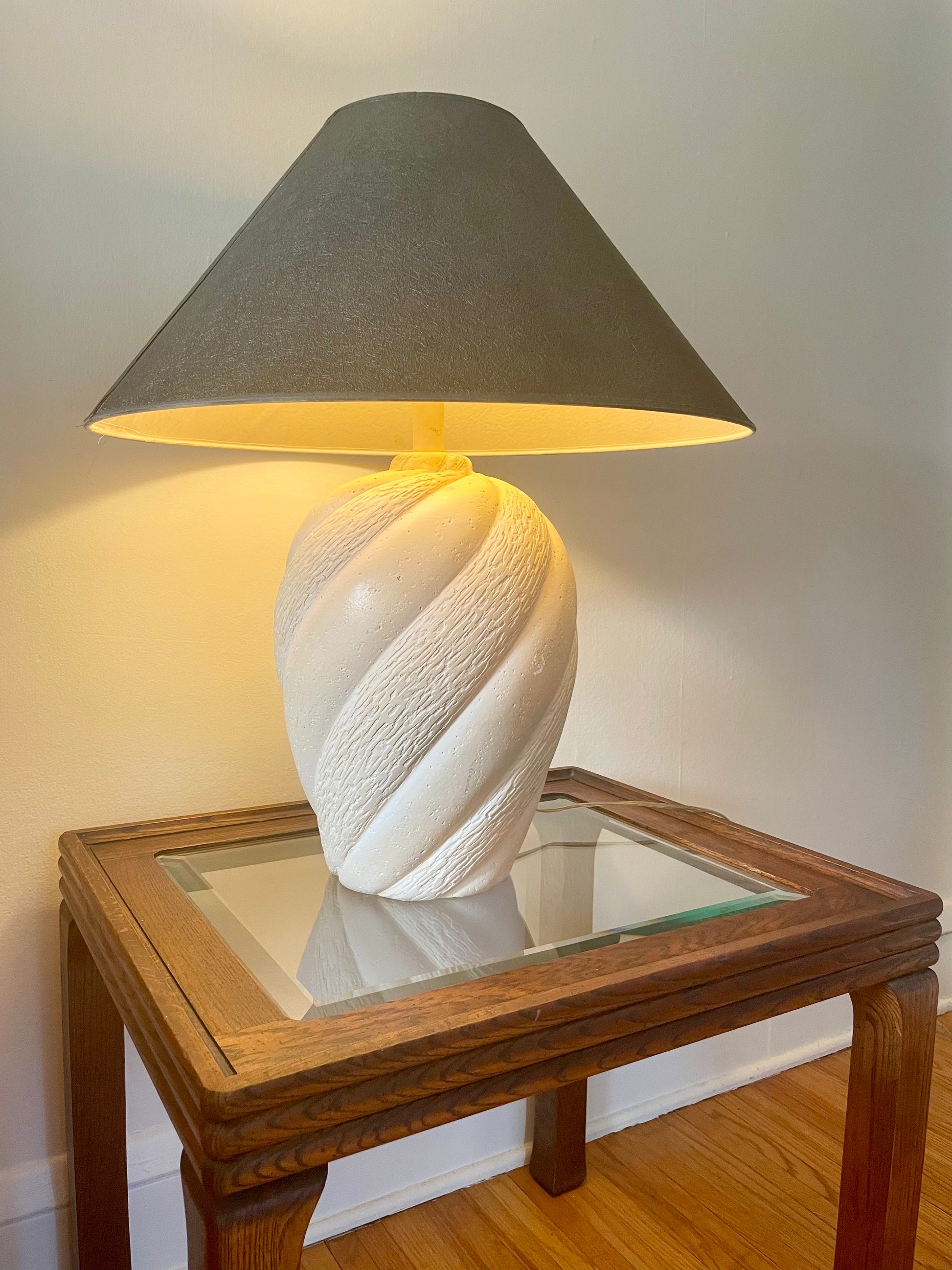 A cream-colored plaster table lamp base with a textured spiral design shown with a graphite tapered lampshade and illuminated in a warm yellow glow, sitting on a glass-top wood side table in front of a plain wall.