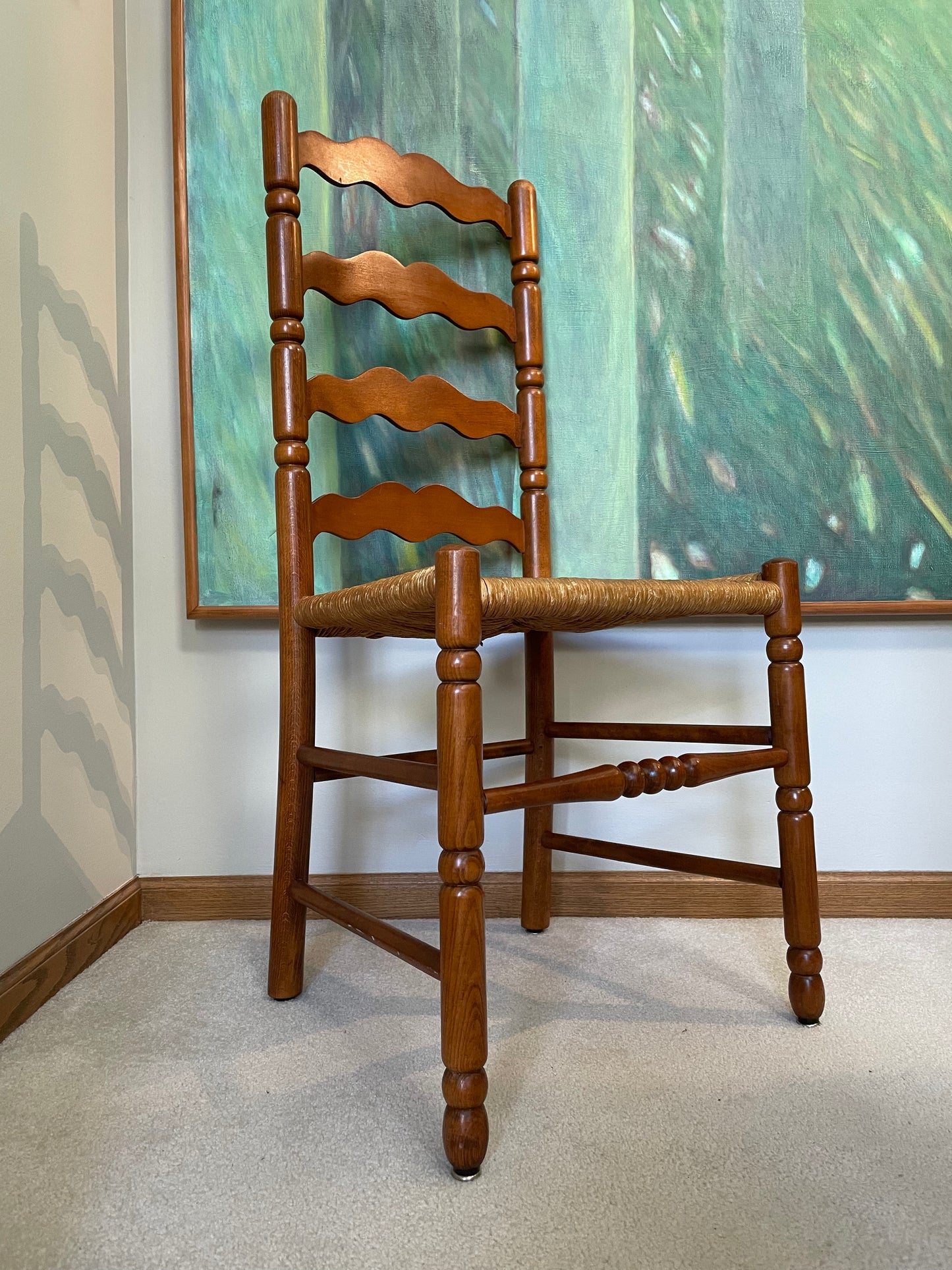 A wooden ladder-back dining chair with a woven seat, positioned in front of a large green and teal framed painting on a white wall.