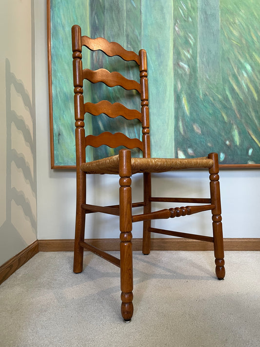 A wooden ladder-back dining chair with a woven seat, positioned in front of a large green and teal framed painting on a white wall.