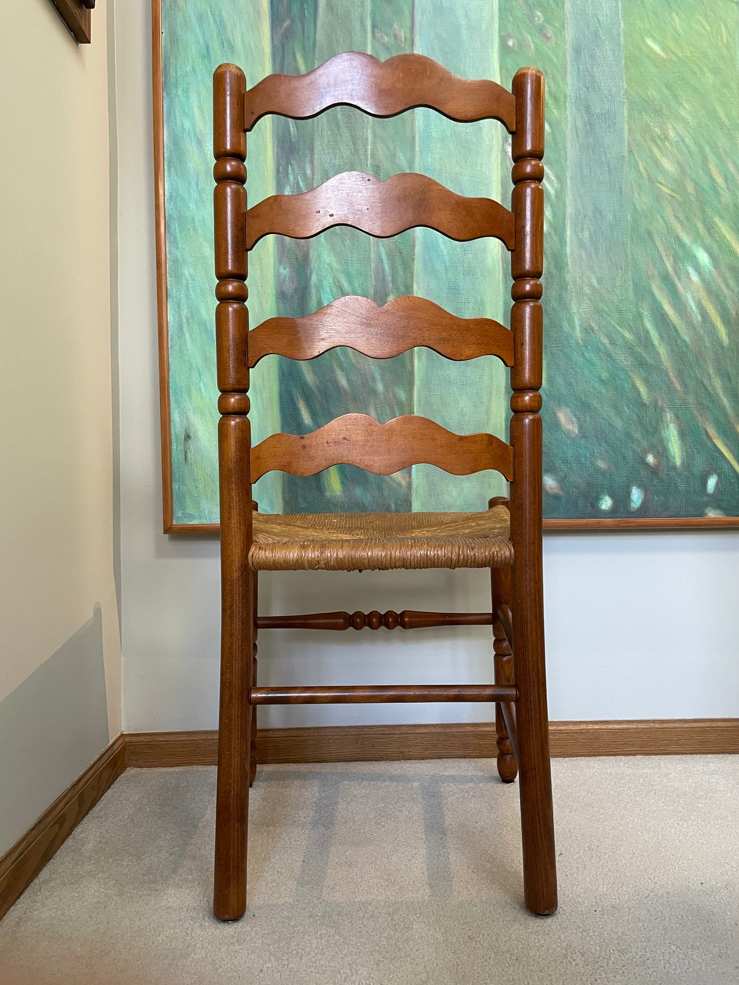 Backside view of a wooden ladder-back dining chair with a woven rush seat, positioned in front of a framed green landscape painting on a white wall.