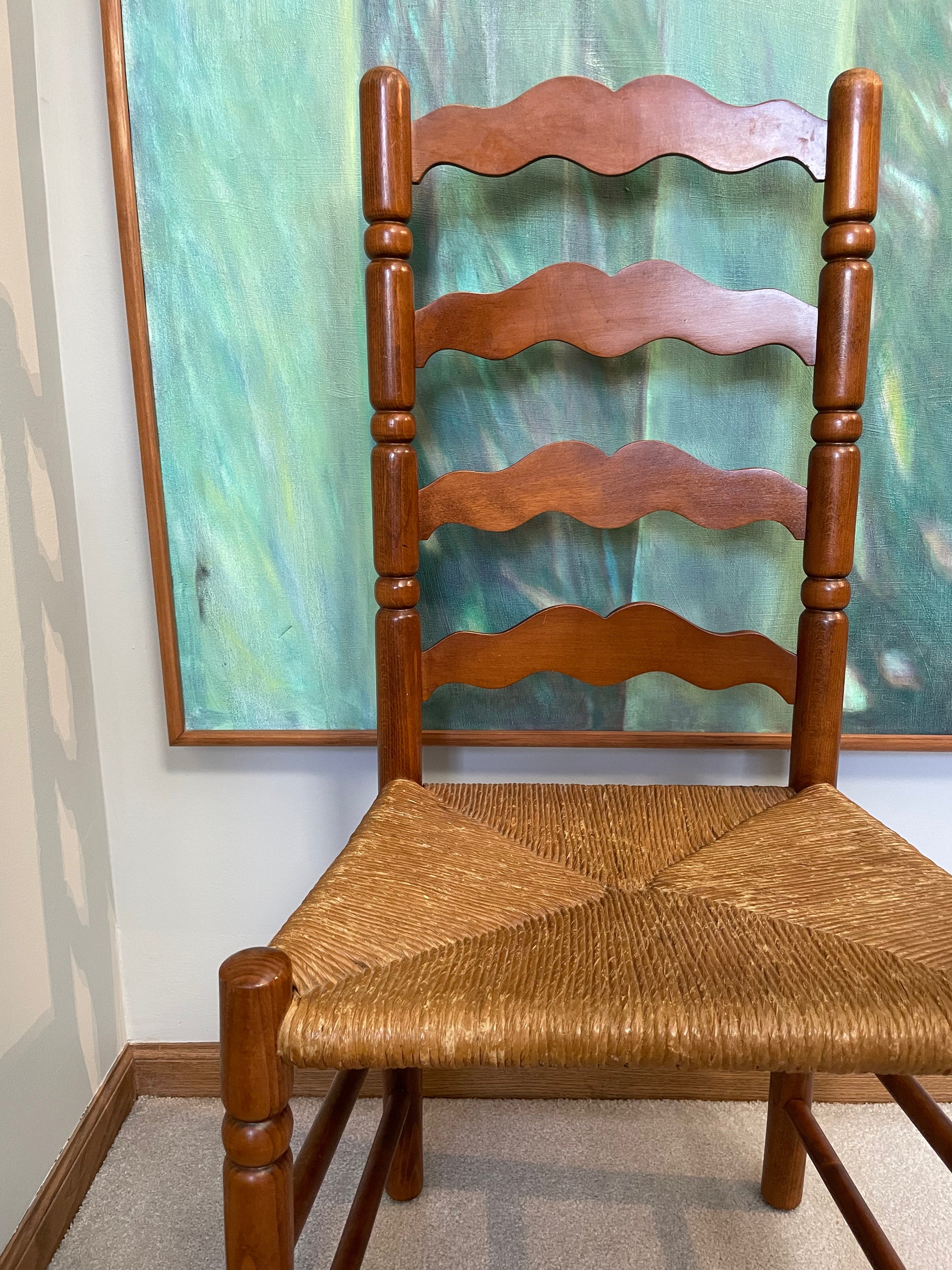 A wooden ladder-back dining chair with a woven rush seat, positioned in front of a large framed abstract painting.