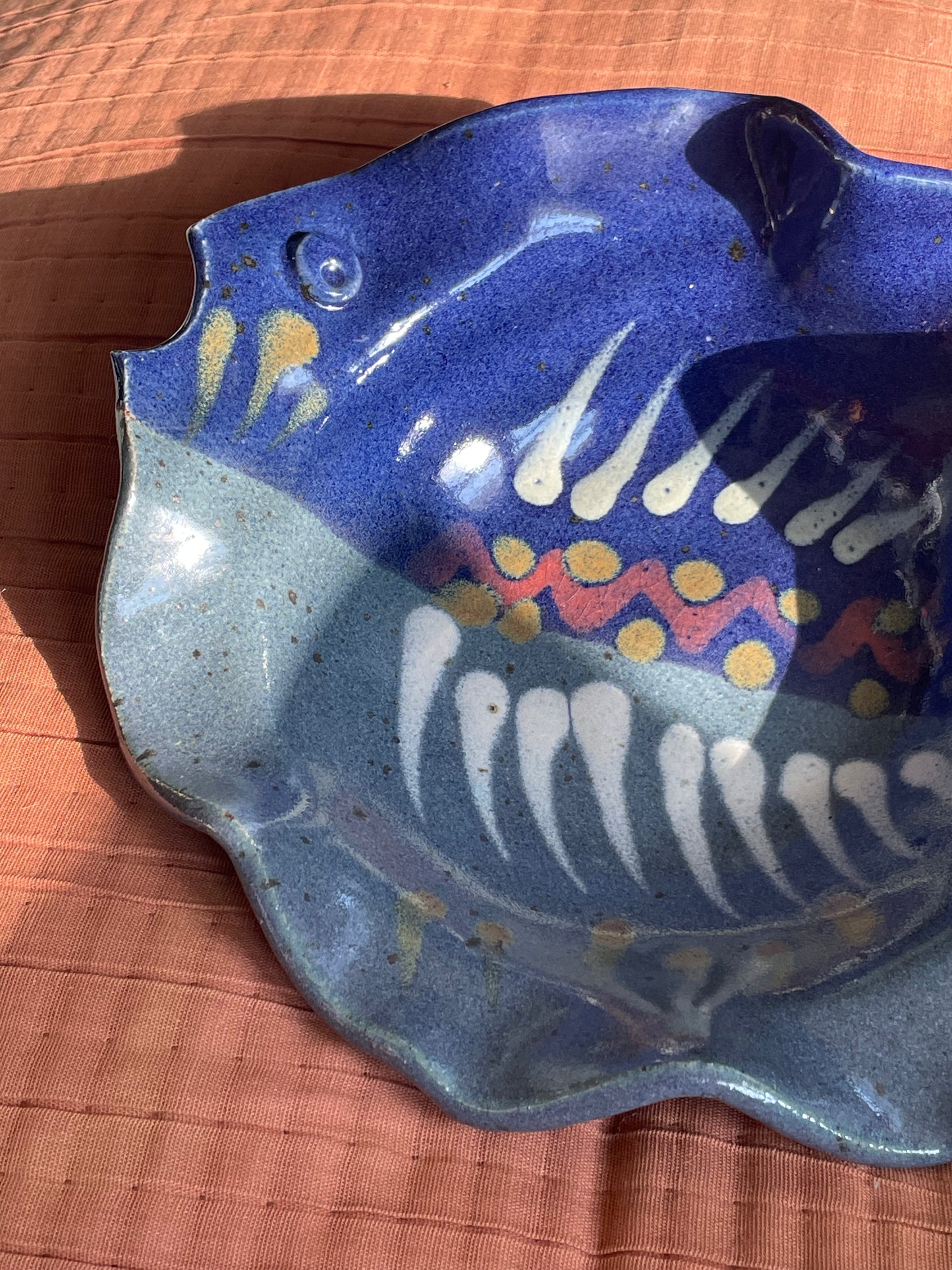 A handmade ceramic fish-shaped dish with a blue and gray speckled glaze, decorated with white teeth-like patterns and yellow and red accents, resting on a textured terracotta surface.