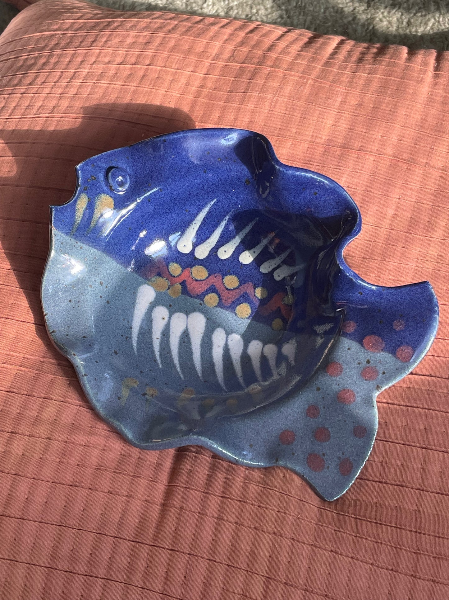 A handmade ceramic fish-shaped dish with a blue and gray speckled glaze, decorated with white teeth-like patterns and yellow and red accents, resting on a textured terracotta surface.
