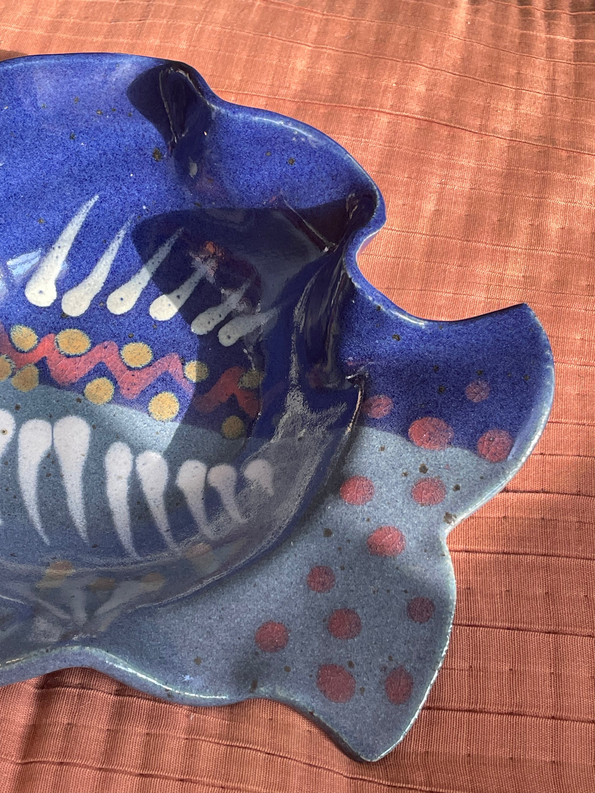 A close-up of the tail of a handcrafted ceramic fish-shaped dish with a glossy blue and light blue glaze, decorated with white stripe details, red and yellow polka dots, and pink spots, set against a textured terracotta background.