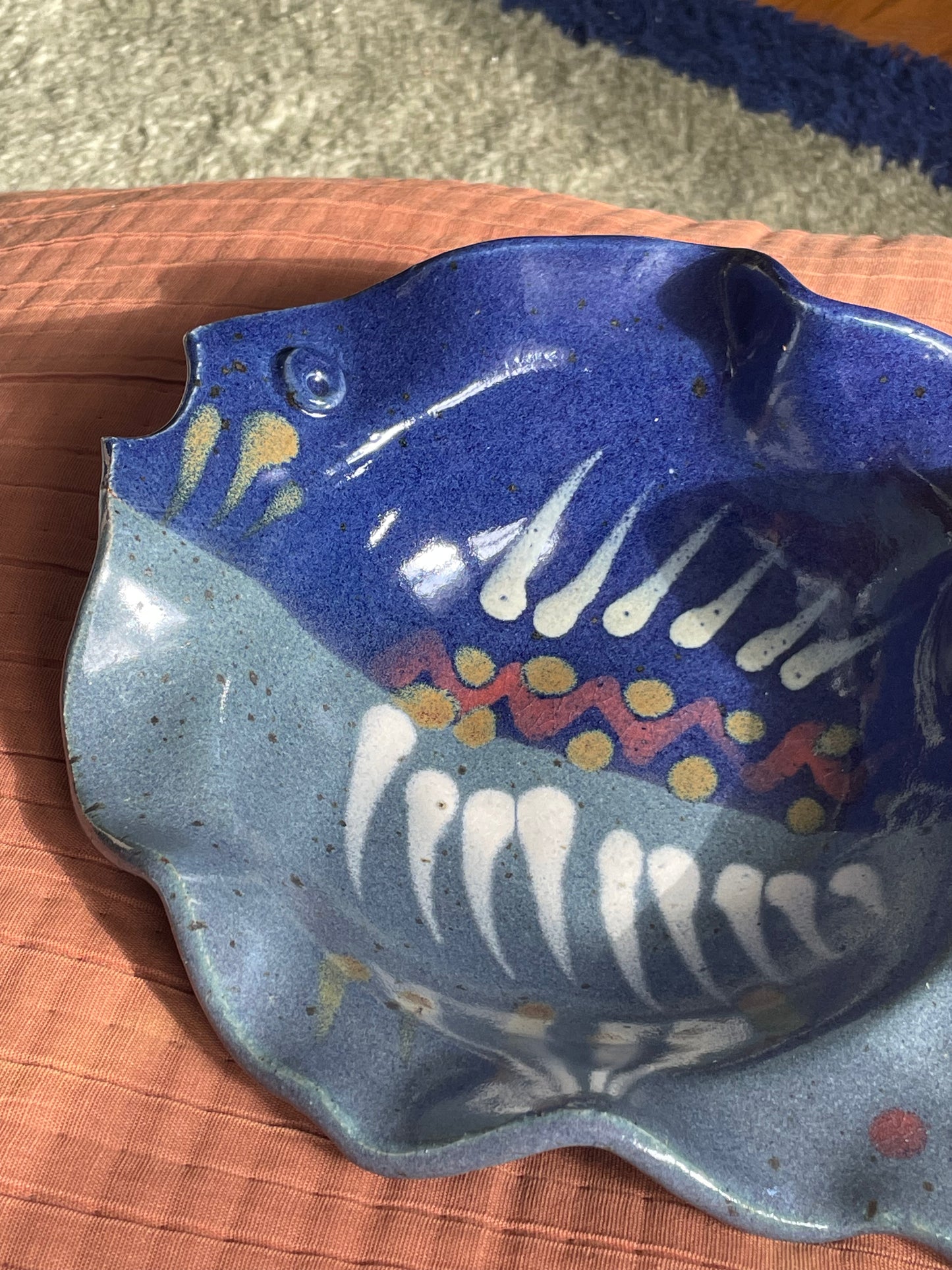 A handmade ceramic fish-shaped dish with a blue and gray speckled glaze, decorated with white teeth-like patterns and yellow and red accents, resting on a textured terracotta surface.