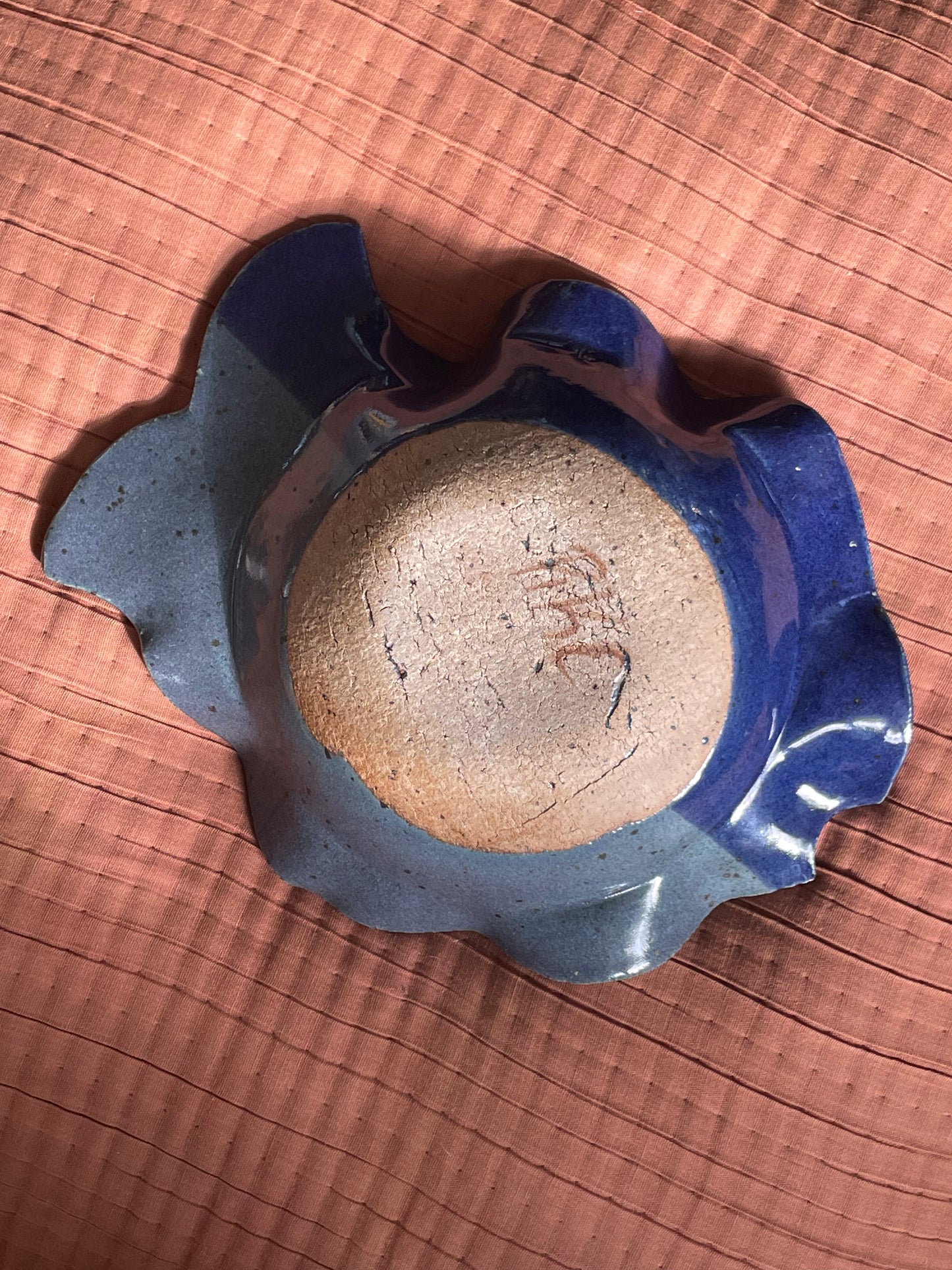 The underside of a handmade ceramic fish-shaped dish with a scalloped, wavy rim, featuring glossy deep blue glazed edges and raw unglazed clay base engraved with ‘RKC’. The bowl is photographed from a top-down perspective on a textured terracotta-colored surface.
