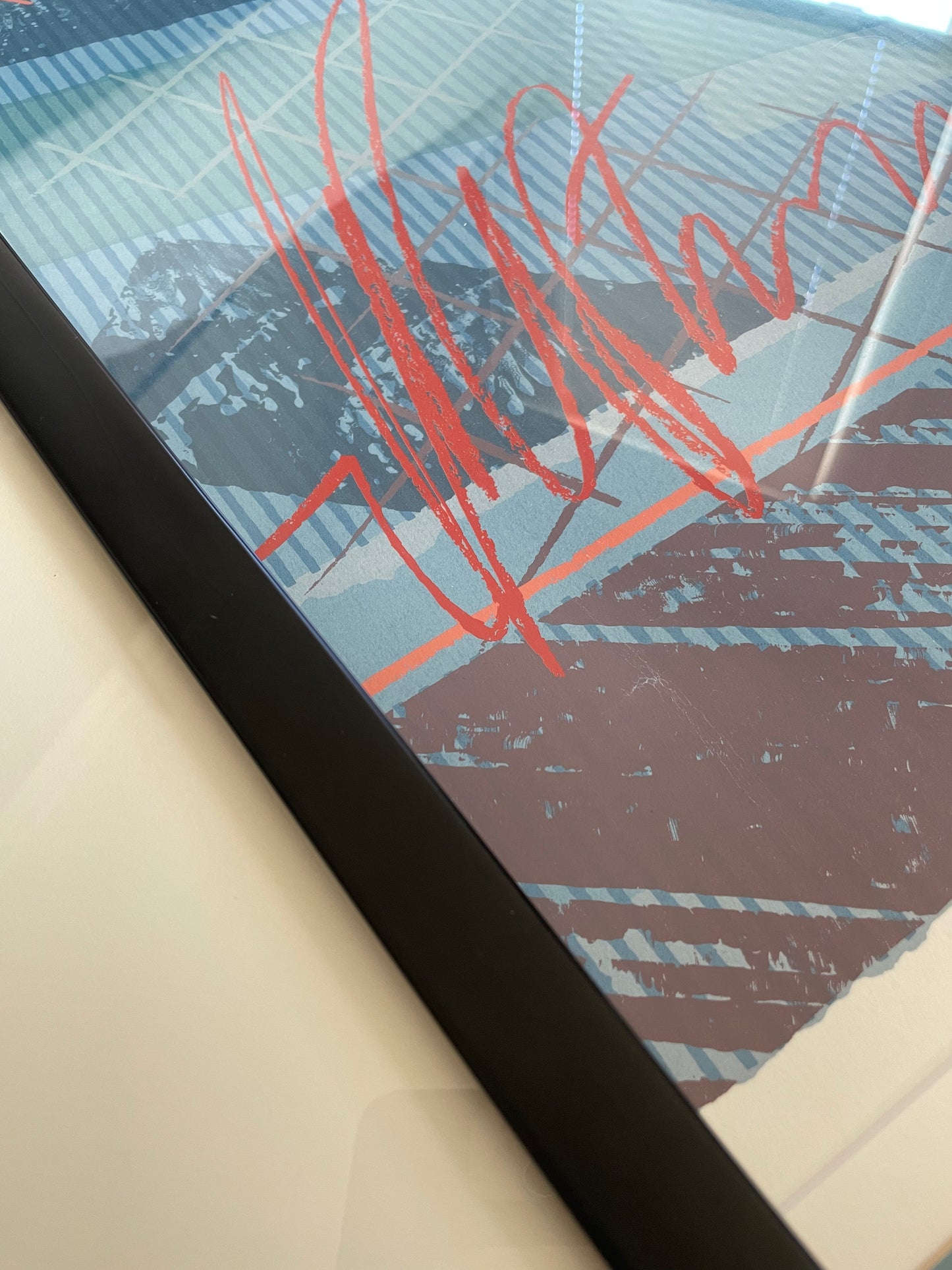 A close-up of a framed art print featuring an abstract design with red scribbled lines over a background of blue, red, and mauve geometric shapes.