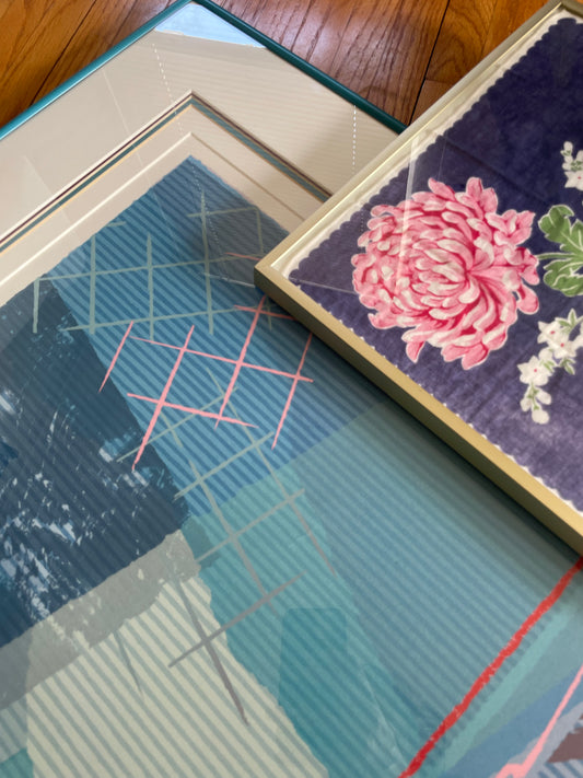 Two framed artworks resting on a wooden floor. The print on the left features a blue and teal geometric pattern with pink grid lines and framed in a teal frame, while on the right a vintage indigo handkerchief with a block-printed pink peony flower and green leaves is shown in a gold frame.