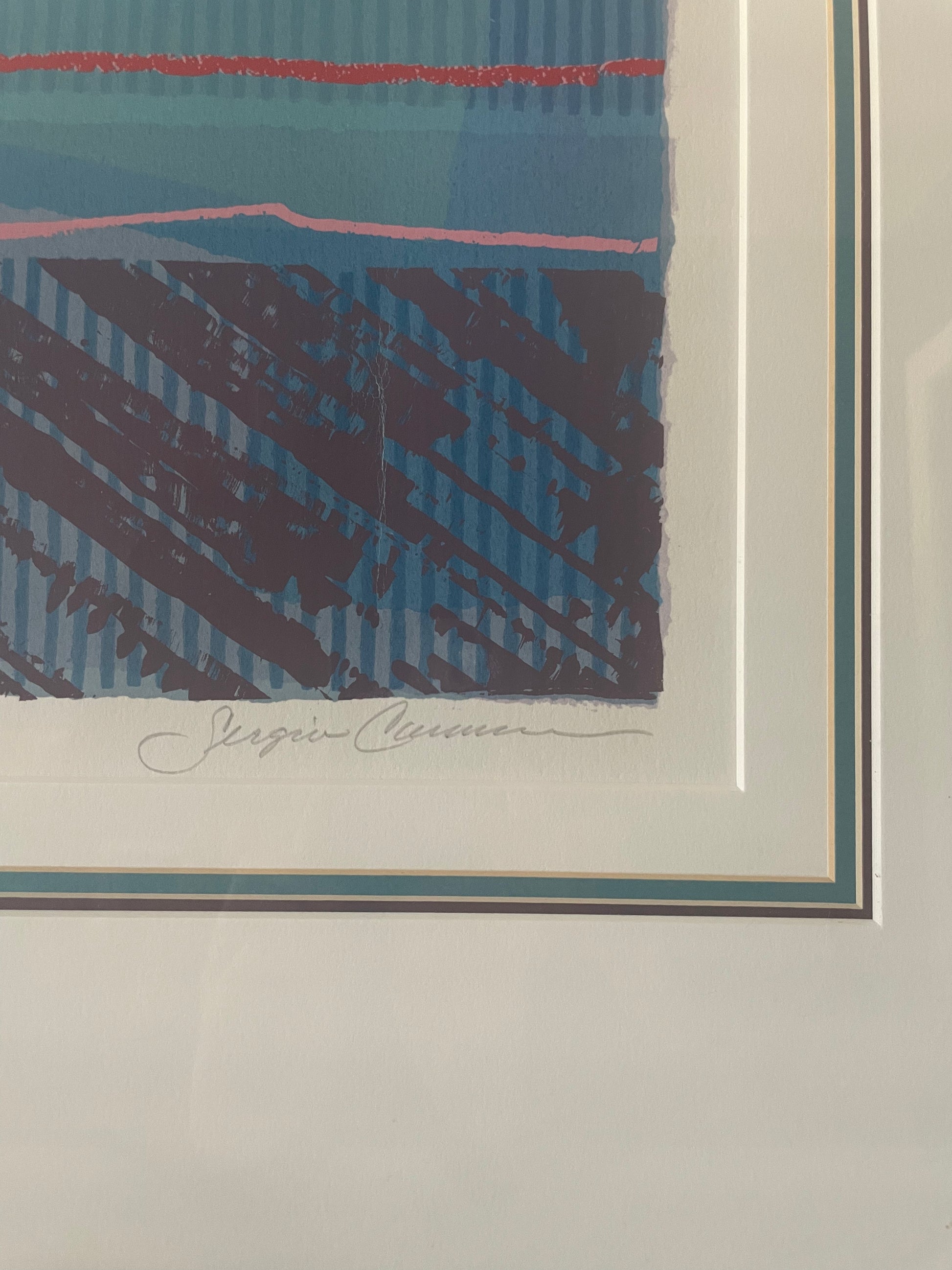 A close-up of a framed abstract art print featuring blue, red, mauve, and pink brushstrokes, with a white mat and a signature below the print.
