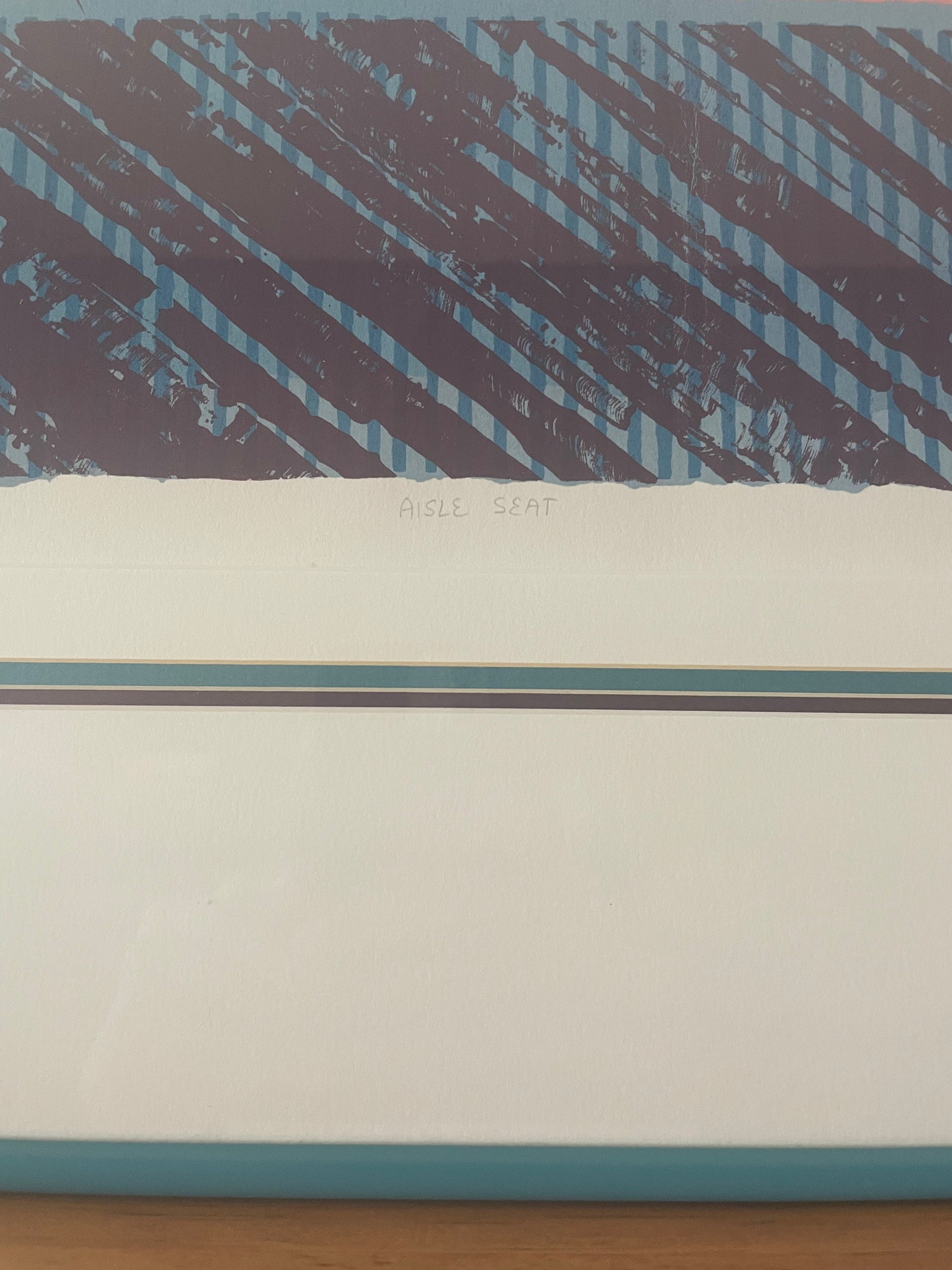 A close-up of the center bottom of an art print showing a geometric design with a dark purple and light blue diagonal striped pattern on the top half, the title of the print ‘Aisle Seat,’ and the layered matting inside a teal frame. 