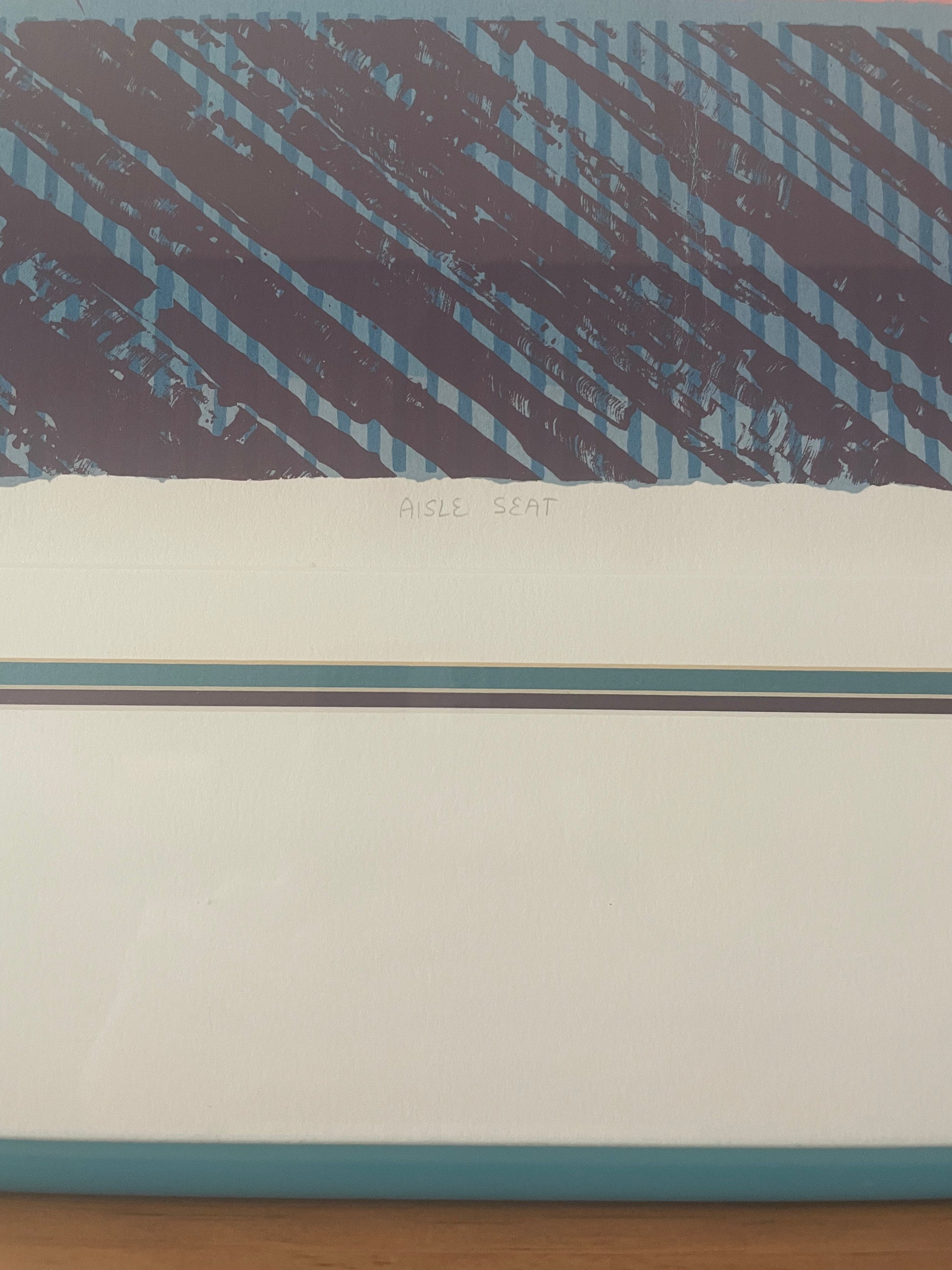A close-up of the center bottom of an art print showing a geometric design with a dark purple and light blue diagonal striped pattern on the top half, the title of the print ‘Aisle Seat,’ and the layered matting inside a teal frame. 