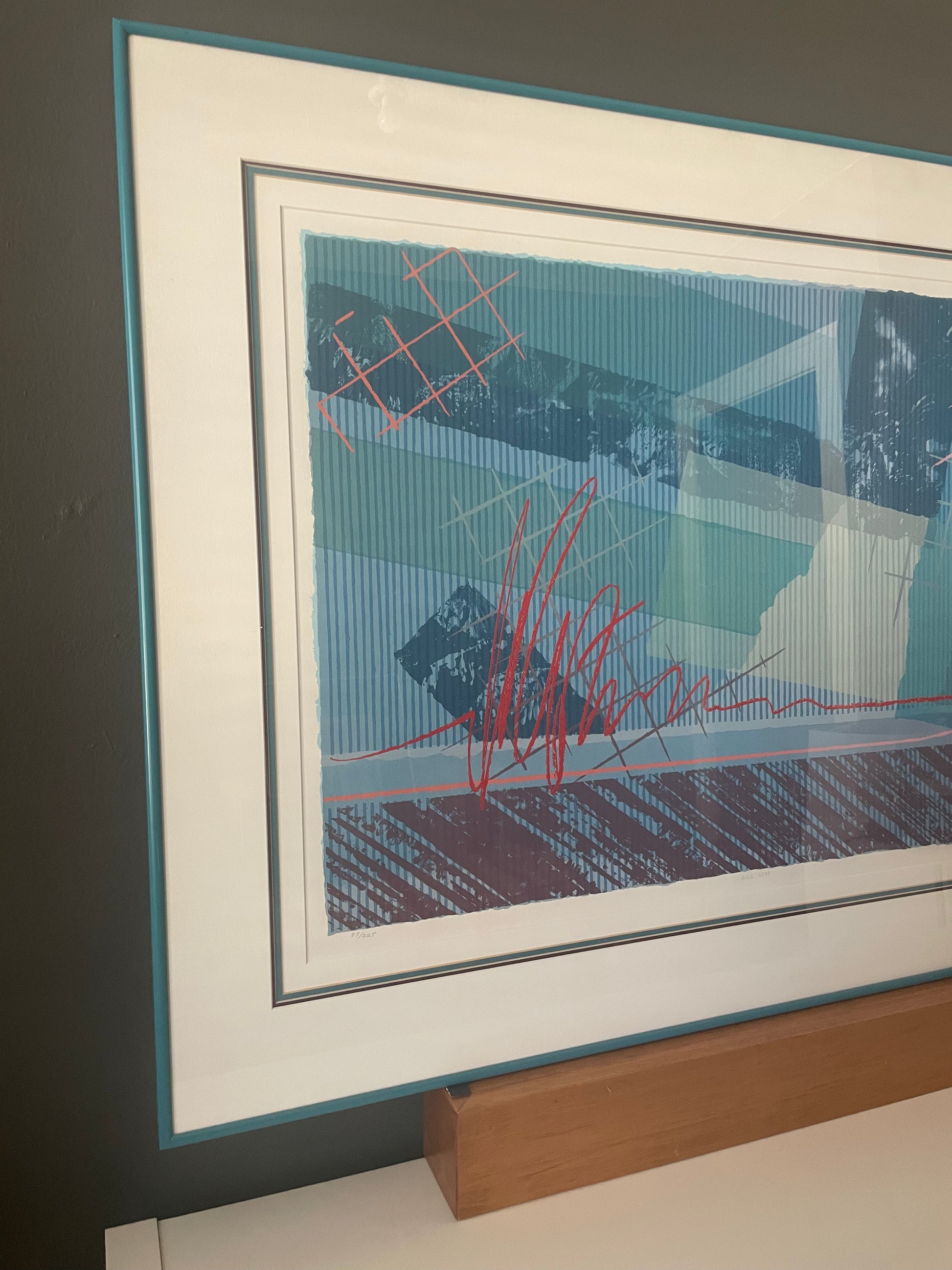 Framed abstract wall art leaning against a dark gray wall above a wooden console. The artwork features a blue and teal background with red sketch-like lines and dark gray geometric shapes, surrounded by a wide white mat and a thin teal frame.