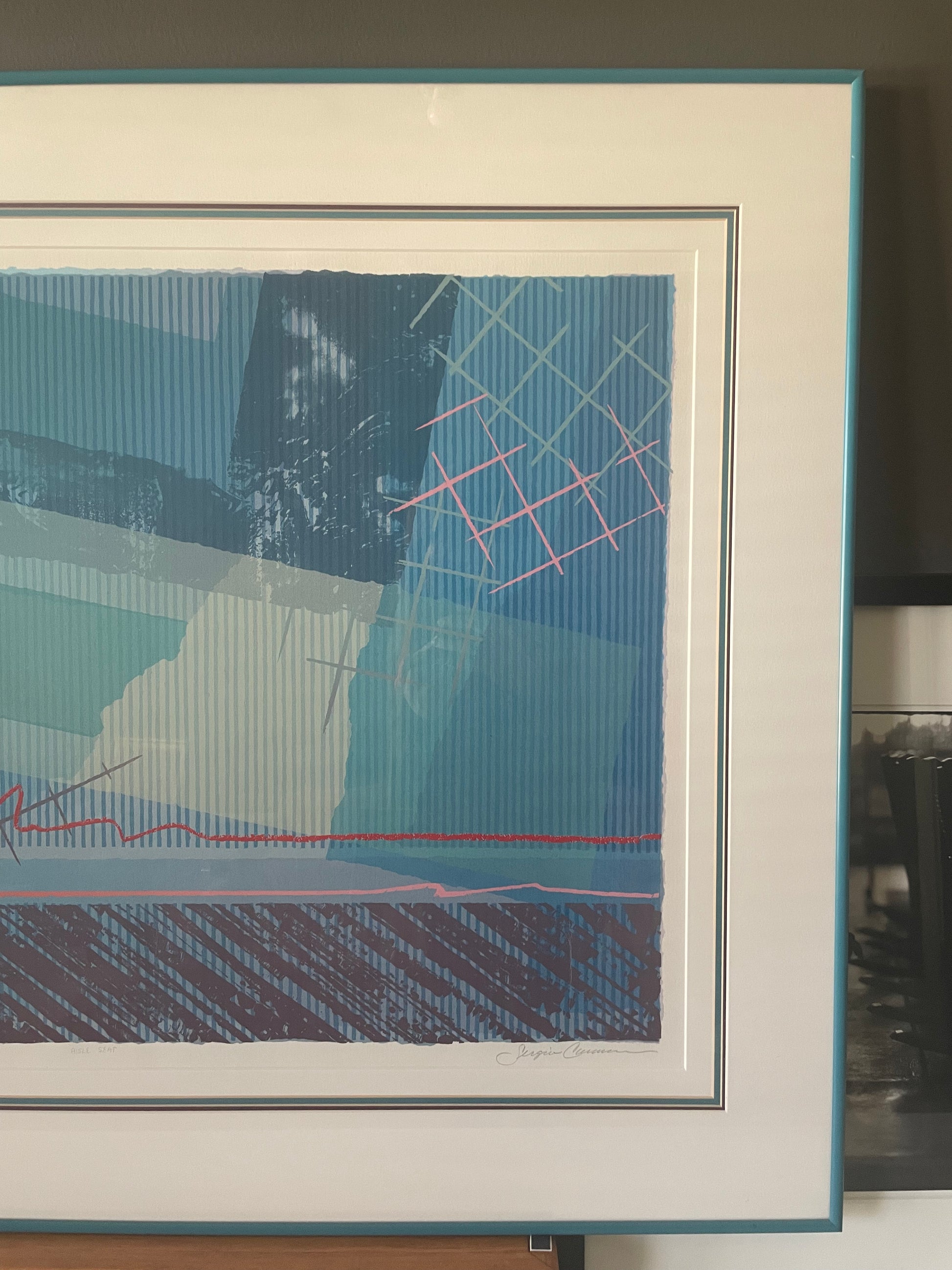 A framed abstract art print featuring geometric shapes and textured brushstrokes in shades of blue, teal, white, and pink, with a wide white mat and a teal frame.