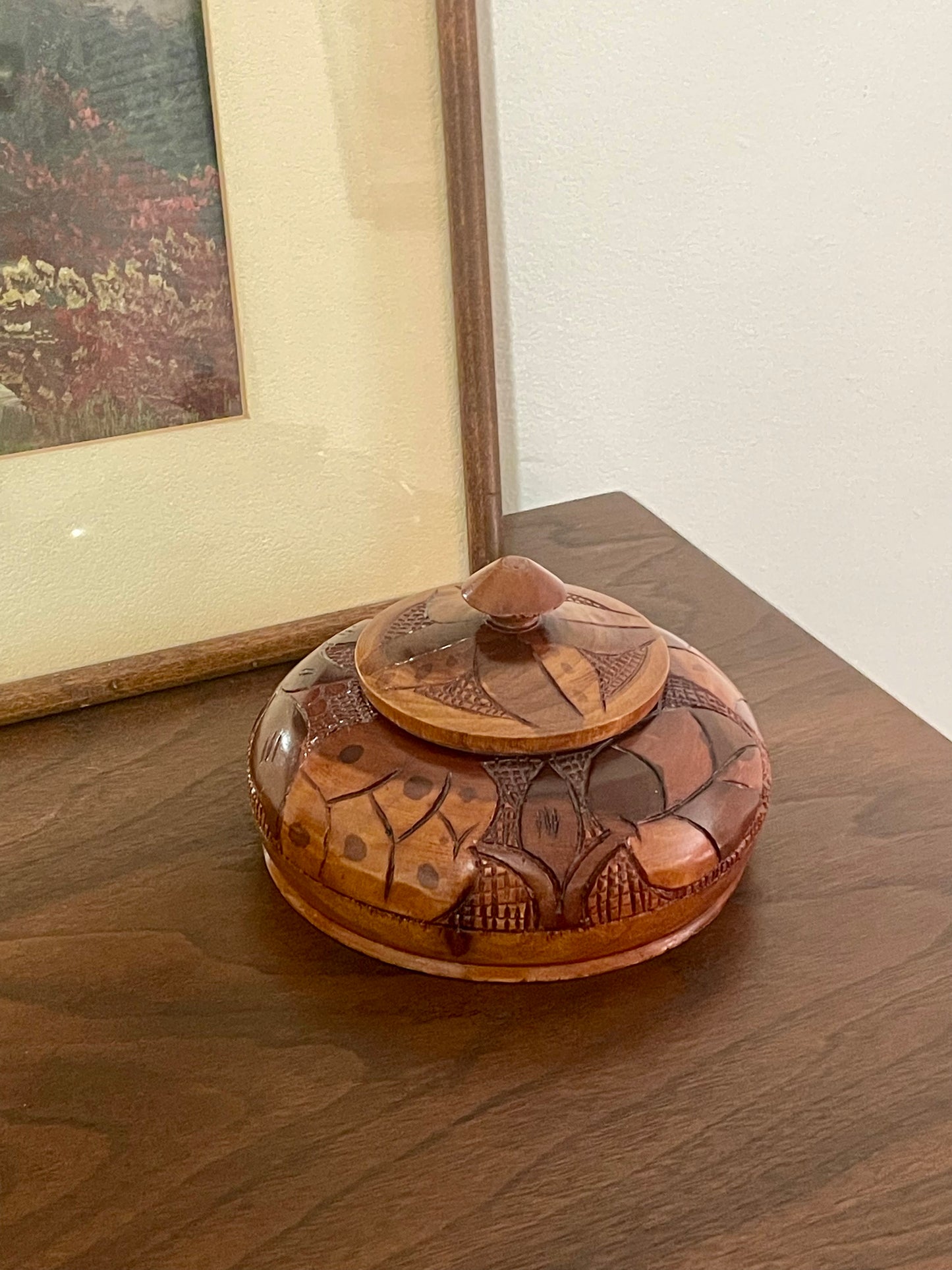 A small carved wooden box with a lid, featuring geometric and floral patterns in natural wood tones, resting on a dark wooden table next to a framed picture.